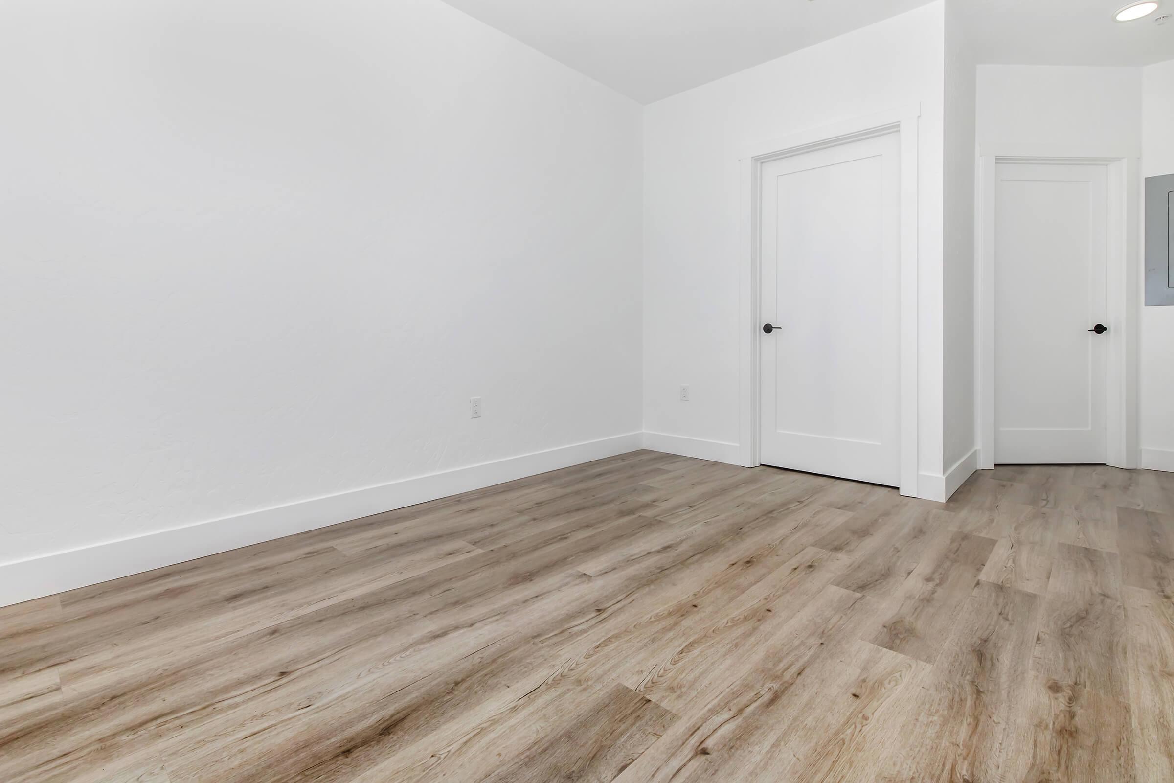 A spacious, empty room with light-colored walls and wood-like flooring. A closed door on the left side leads to another space, while a door on the right side is partially visible. The room is well-lit, creating a clean and minimalistic atmosphere.