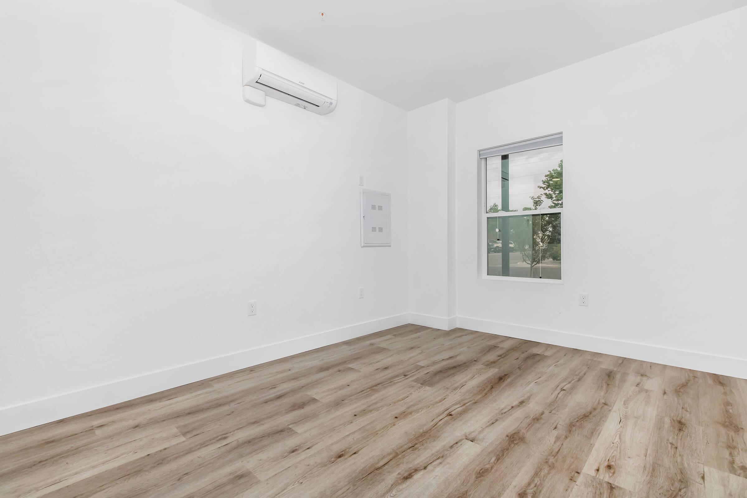 Empty room with light-colored walls and wooden flooring. A window lets in natural light, and there is a small air conditioning unit mounted on the wall. A power panel is visible near the window. The space appears clean and contemporary, making it suitable for various uses.