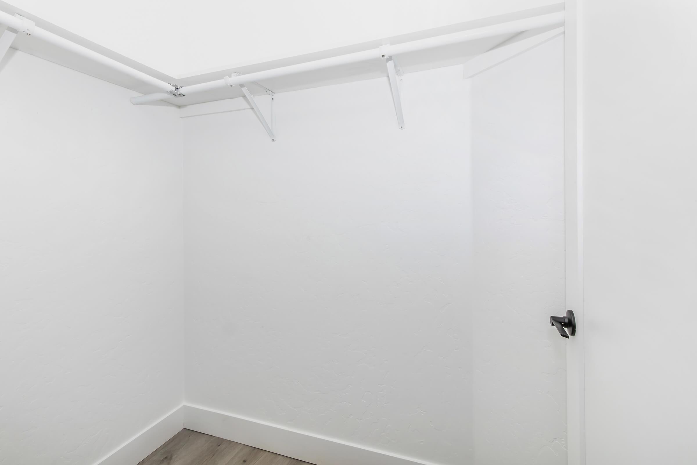 A clean, empty closet with white walls and minimalist design. It features a hanging rod on the left side and a closed door on the right, with a light wooden floor. The space appears bright and uncluttered, suitable for storing clothes or other items.