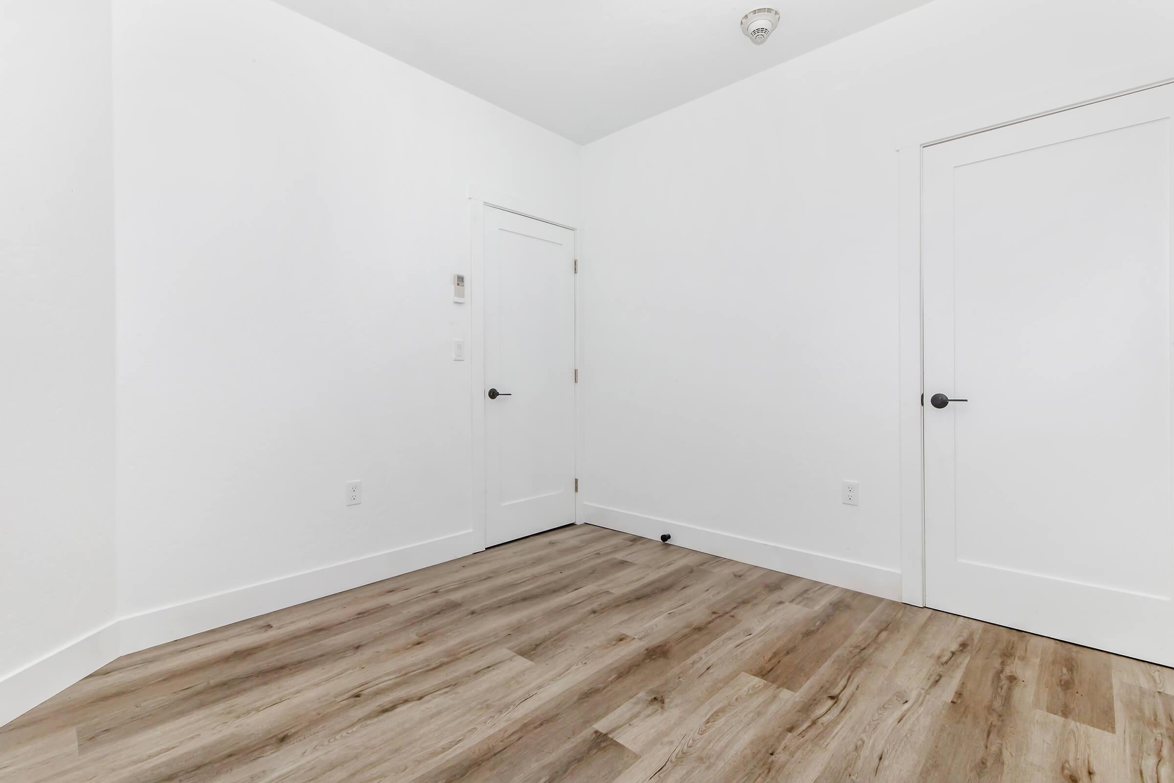 A minimalistic, empty room with white walls and a light wooden floor. There are two doors: one on the left with a black handle and the other on the right, both leading to different spaces. The room is well-lit and devoid of furniture or decorations, creating a spacious and clean appearance.