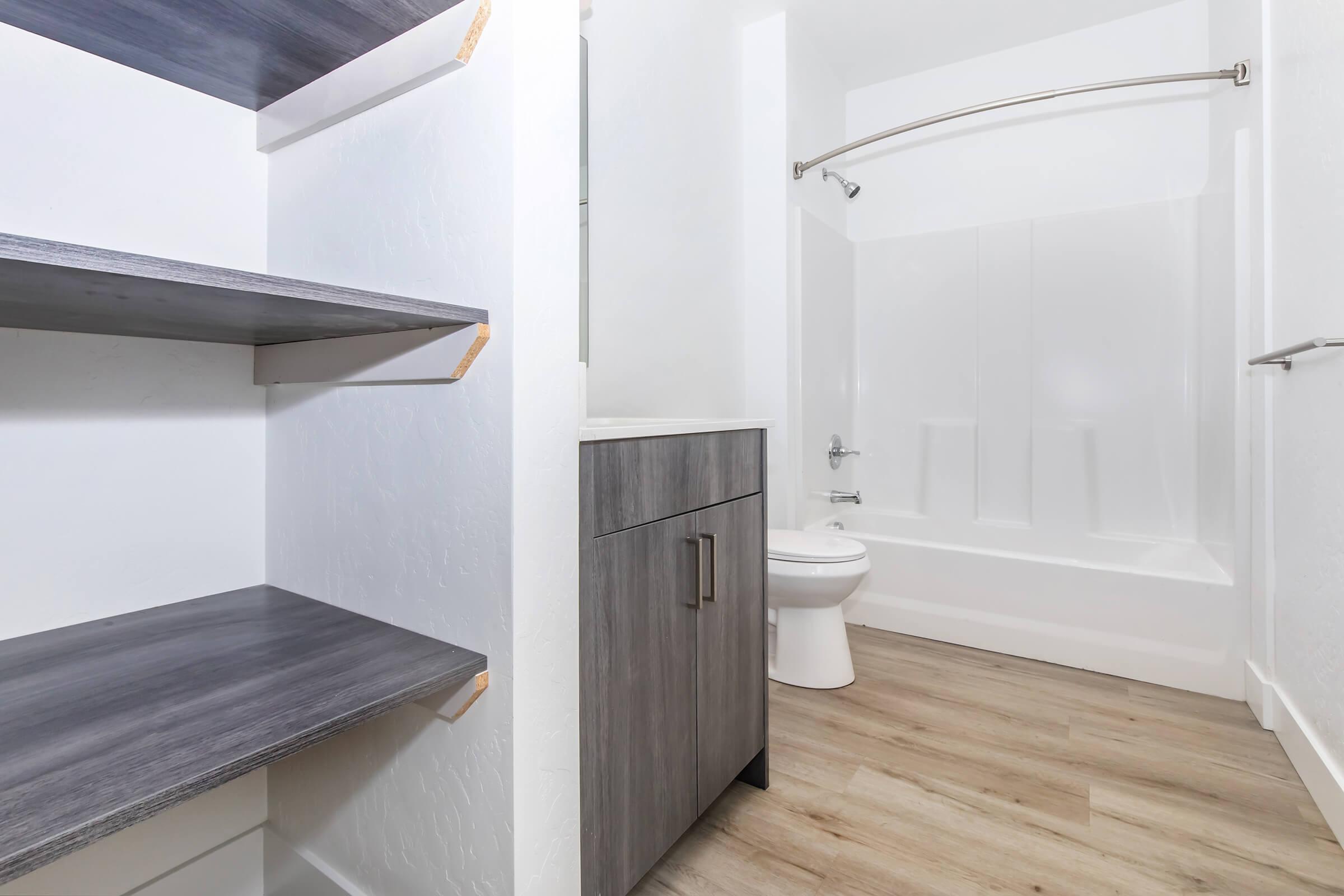 A modern bathroom featuring a shower tub combo, a toilet, and a vanity cabinet with a countertop. To the left, there are open shelves that provide additional storage. The flooring is light-colored wood, and the walls are painted white, creating a clean and bright appearance.