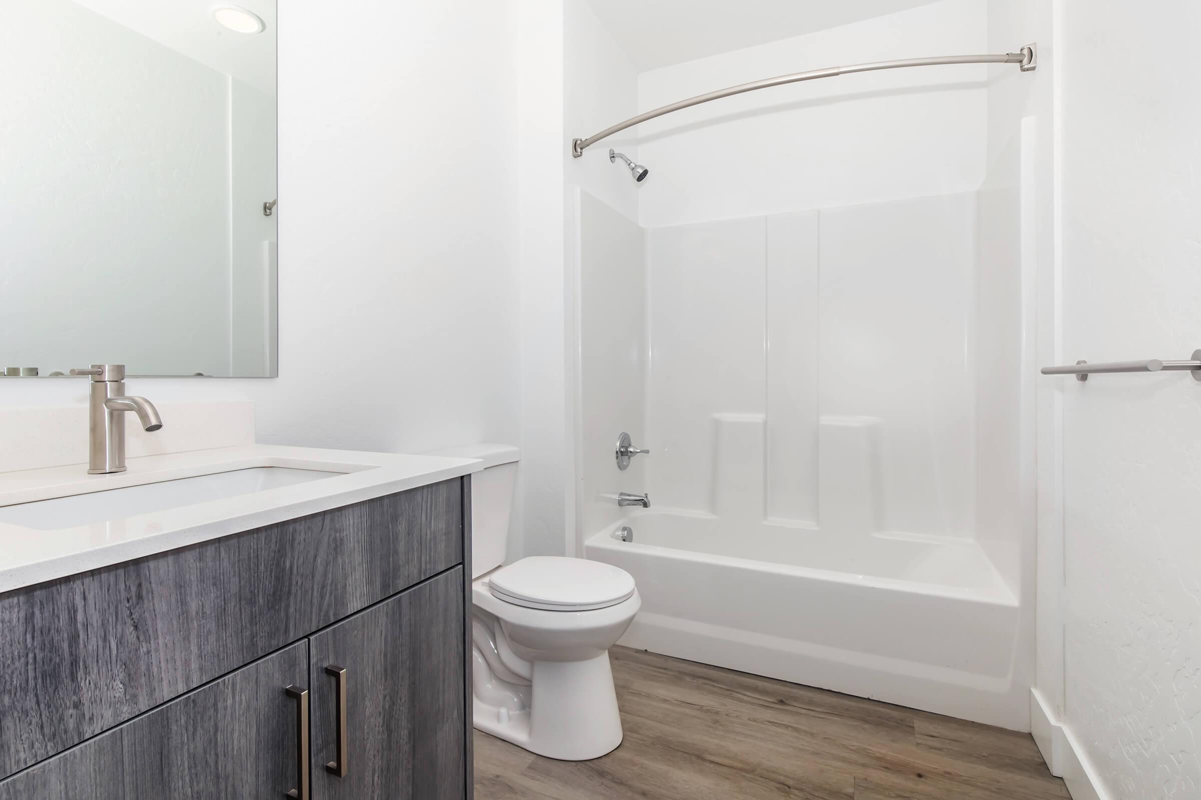 Modern bathroom featuring a white bathtub and shower combo, a sleek vanity with a sink and modern faucet, and a toilet. The walls are painted white, and the floor has light hardwood-like tiles. A mirror is mounted above the sink, enhancing the bright and clean aesthetic of the space.