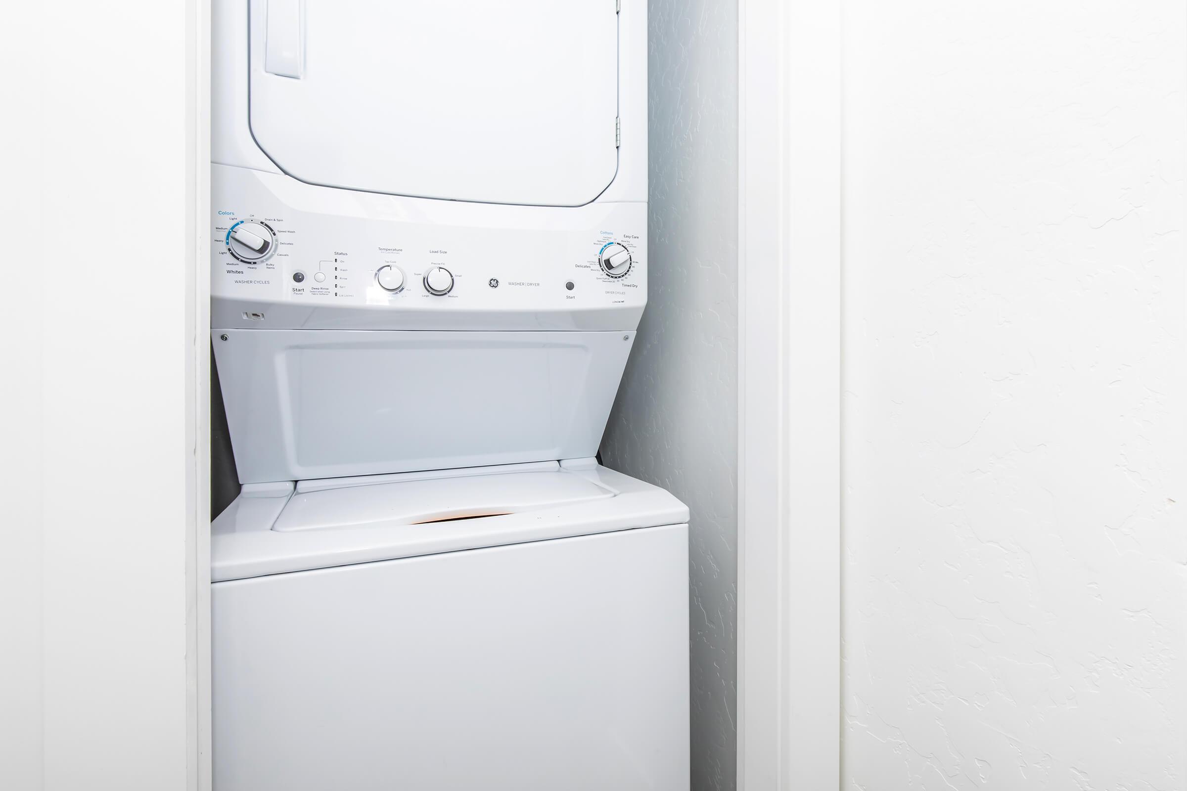 A compact laundry unit featuring a stacked washer and dryer combination, located in a small, well-lit, white space with smooth walls. The appliances are white with control knobs and panels visible, indicating their functionality.