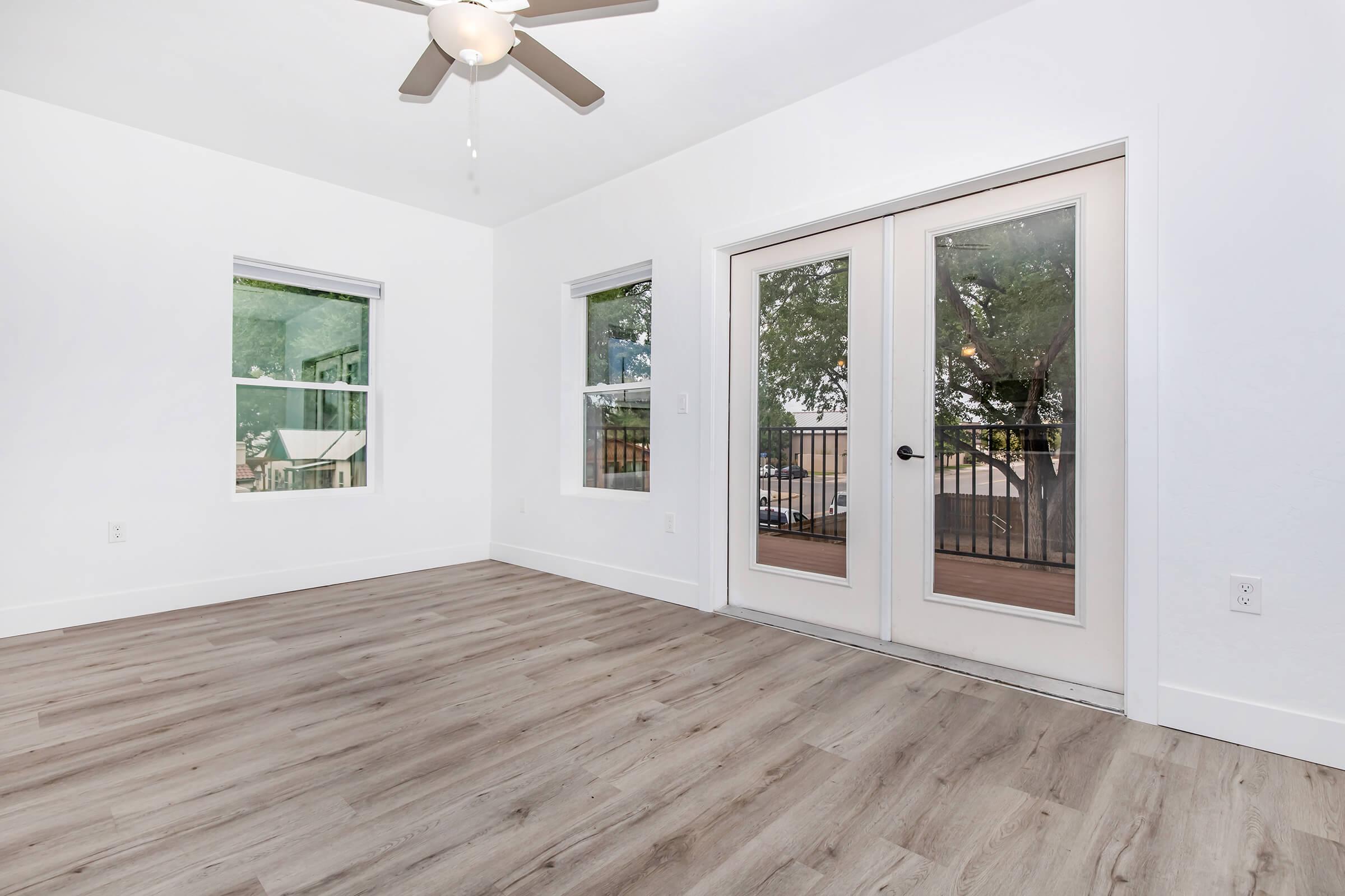 A bright, empty room featuring light-colored walls, large windows with natural light, and a ceiling fan. The flooring is wood-like laminate. French doors lead to a balcony with outdoor views, enhancing the space's openness and potential for design.