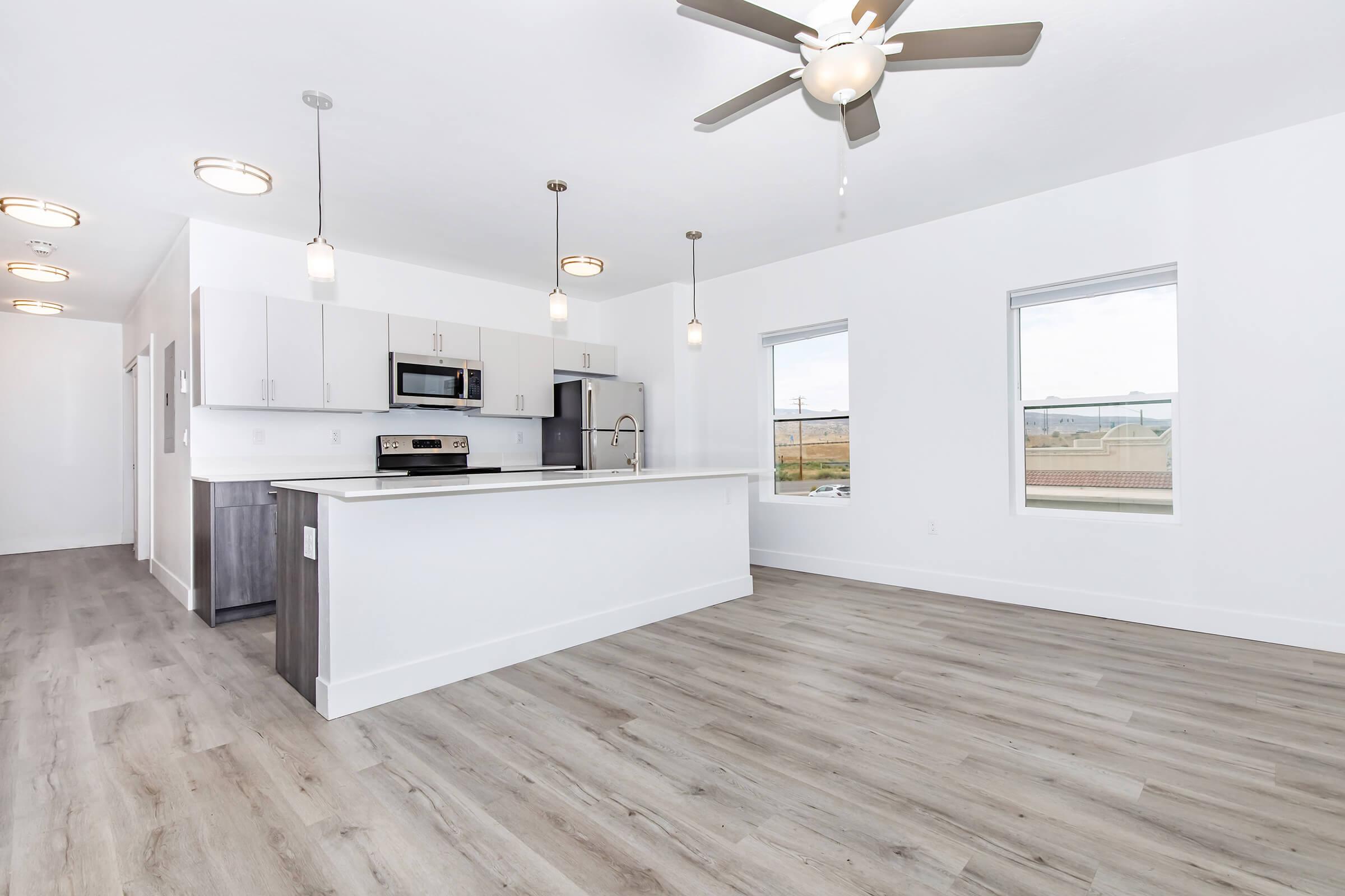 A modern, open-concept kitchen and living area featuring light-colored wood flooring, white walls, and ample natural light from large windows. The kitchen includes sleek cabinetry, stainless steel appliances, and a central island with pendant lighting. A ceiling fan adds to the airy atmosphere.