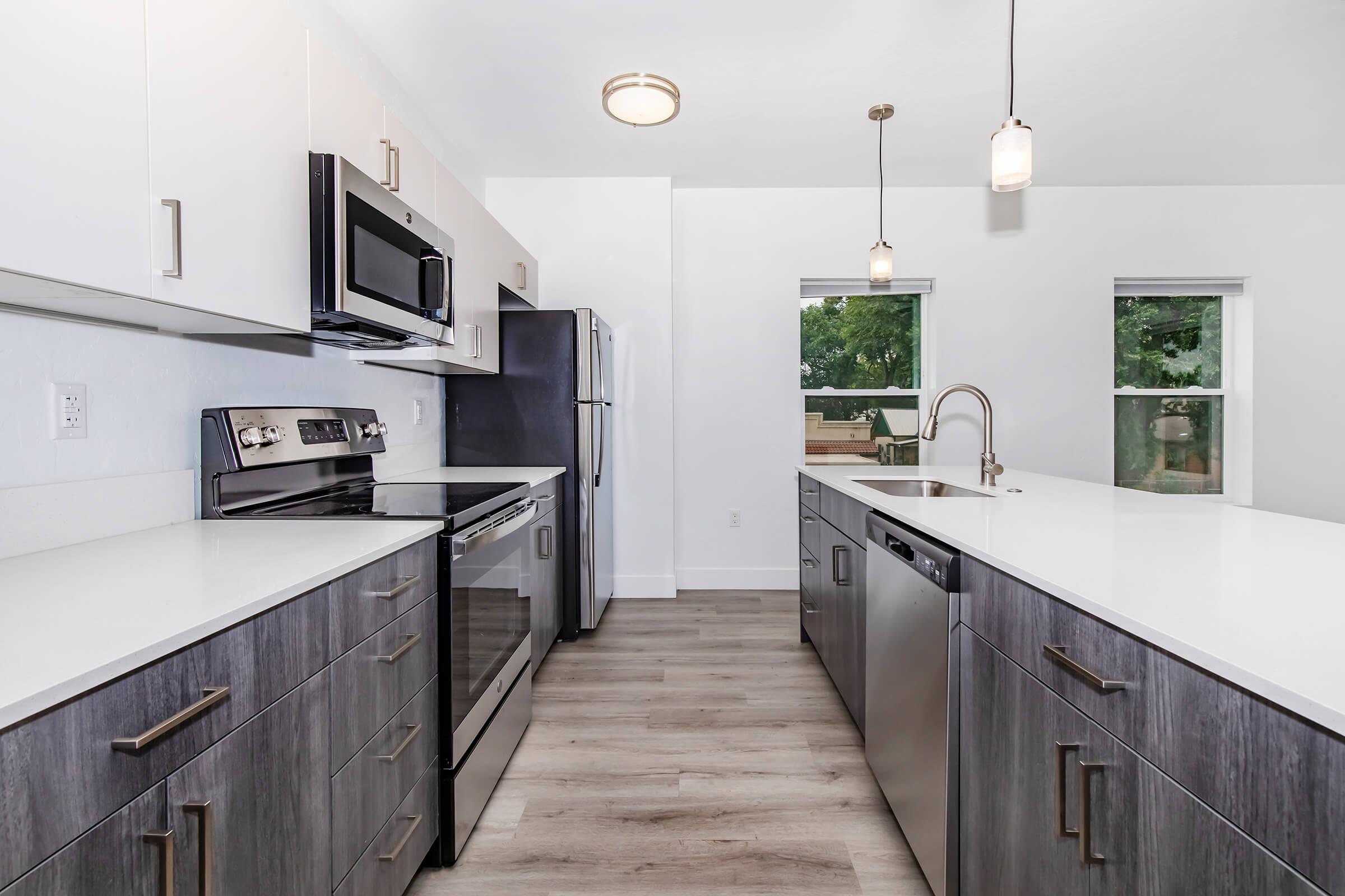 A modern kitchen featuring white and gray cabinets, stainless steel appliances including an oven, microwave, and dishwasher, and a large countertop. The space is well-lit with pendant lights and natural light coming through the two windows. Light-colored wooden flooring completes the contemporary look.