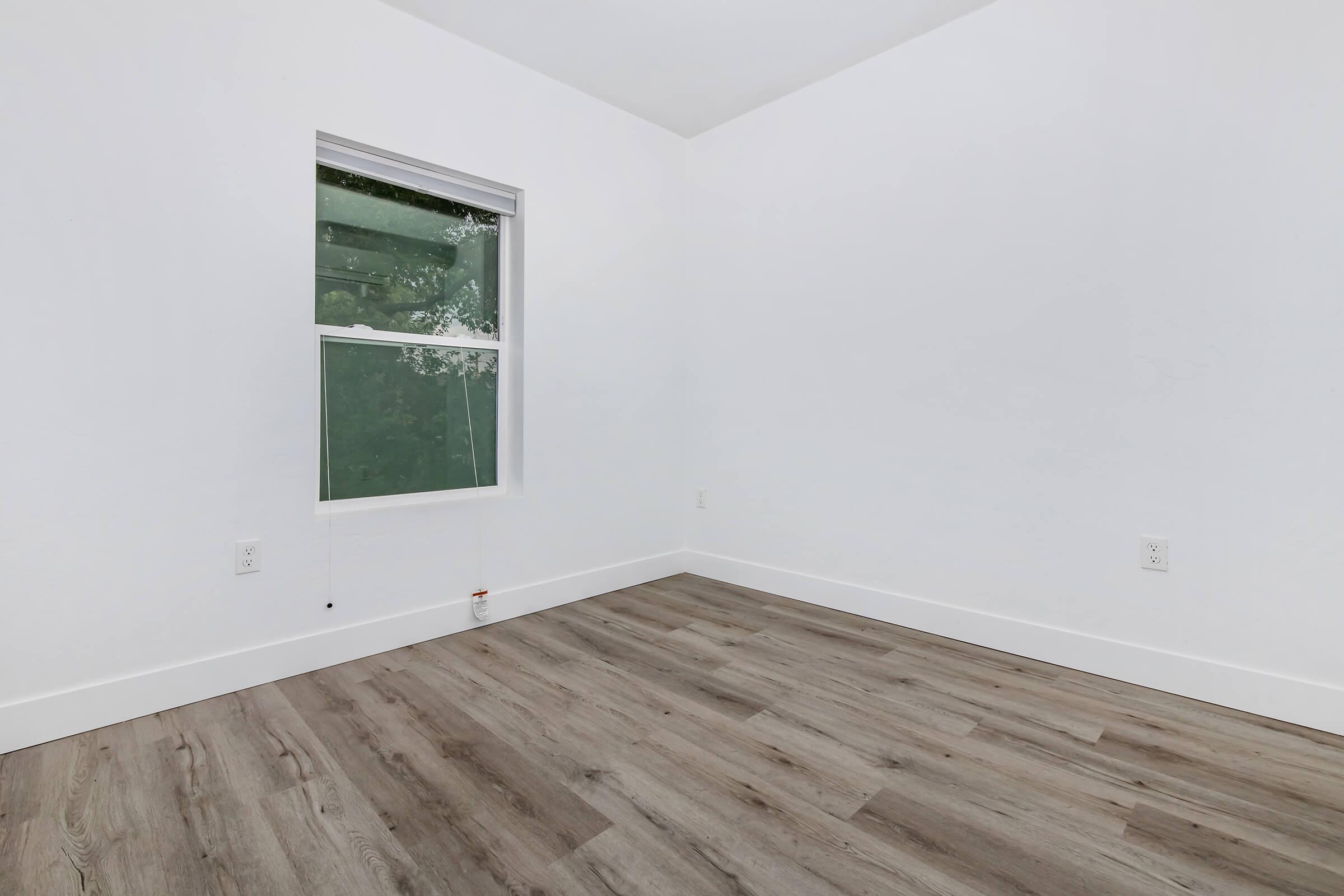 A minimally furnished room featuring white walls and a large window. The floor is made of light-colored wood. The space is empty and bright, with natural light coming through the window, creating a clean and open atmosphere.