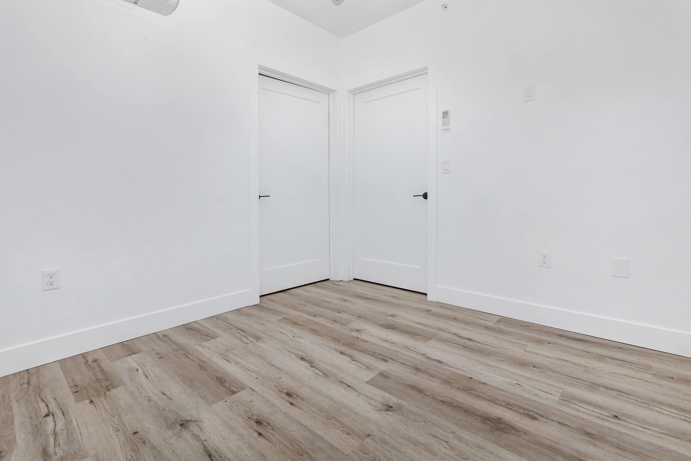 A spacious, empty room with light-colored walls and two closed white doors on the left side. The floor features light wood-like planks, contributing to a clean and modern appearance. The room has a bright and minimalist feel, with no furniture or decorations present.