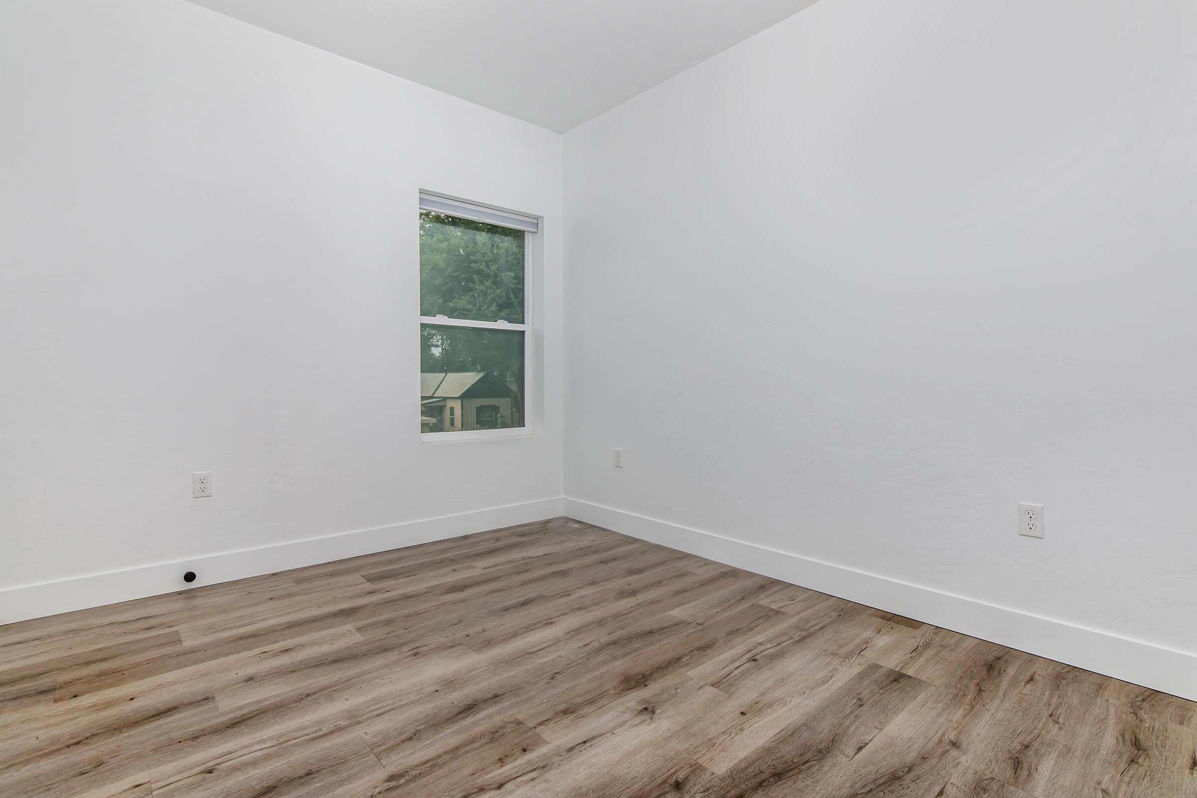 A bright, empty room with white walls and a large window allowing natural light. The flooring is light brown wood laminate. There are no furnishings, creating a minimalist and spacious feel. The window offers a view of the outside greenery.