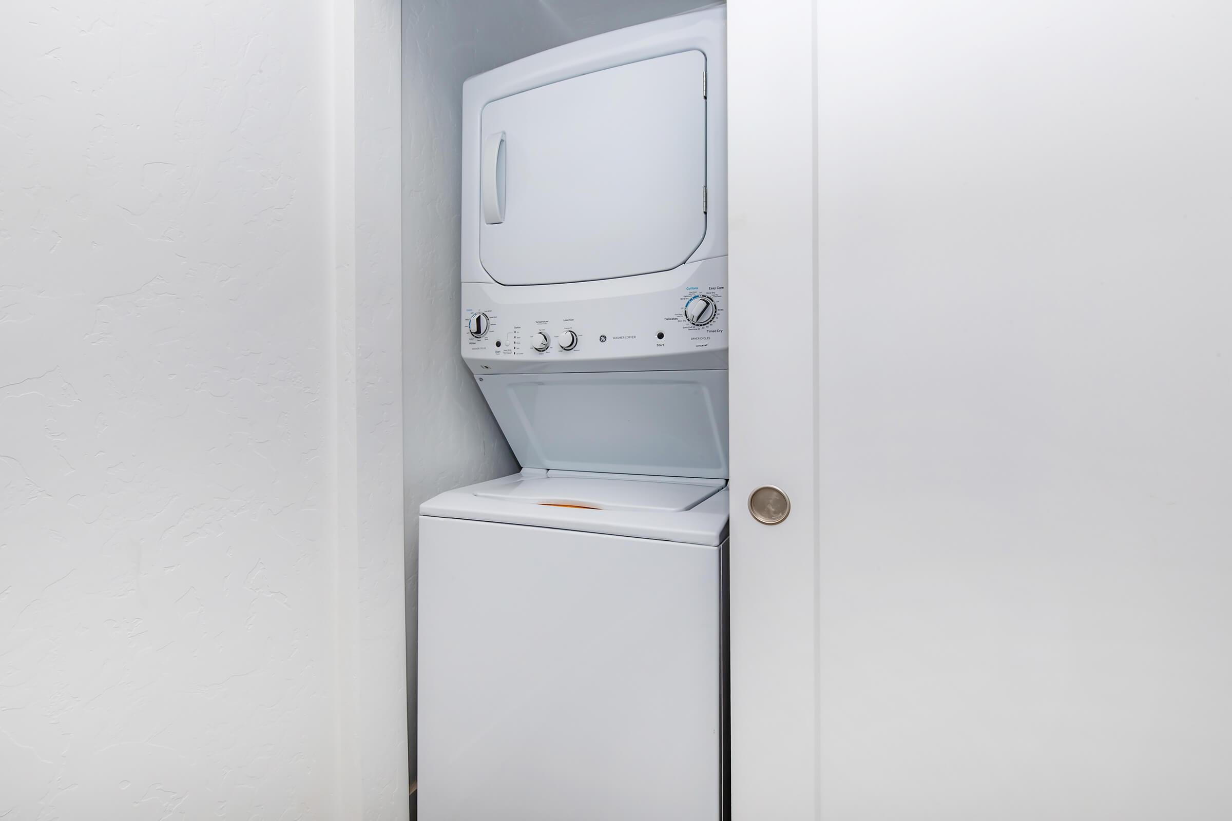 A stacked washer and dryer unit located in a small closet. The washer is at the bottom with a front-loading door, and the dryer, above, has a top-loading door. The space is minimalistic with white walls and a simple door handle.