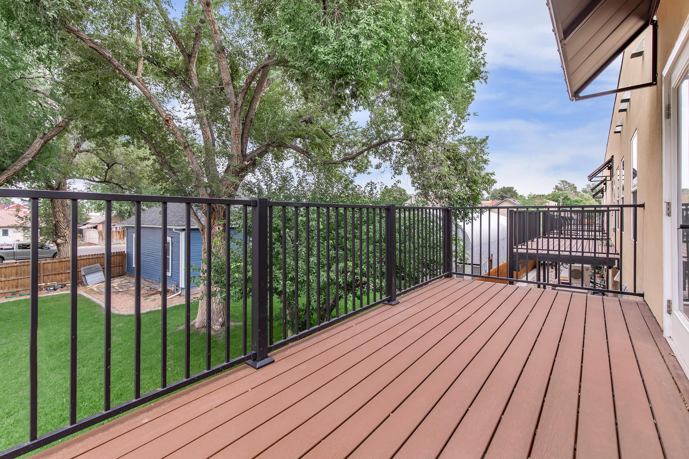 View from a balcony featuring a wooden deck, surrounded by green trees and a well-manicured lawn. In the background, there's a shed and a glimpse of neighboring houses, creating a peaceful residential atmosphere.