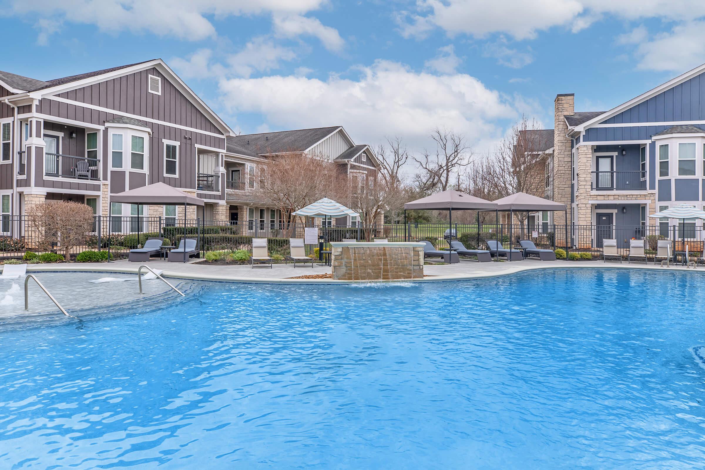 A beautiful outdoor pool area surrounded by comfortable lounge chairs and umbrellas, set against a backdrop of modern residential buildings. The clear blue water reflects the sky, and well-maintained landscaping adds a welcoming touch to the serene atmosphere.