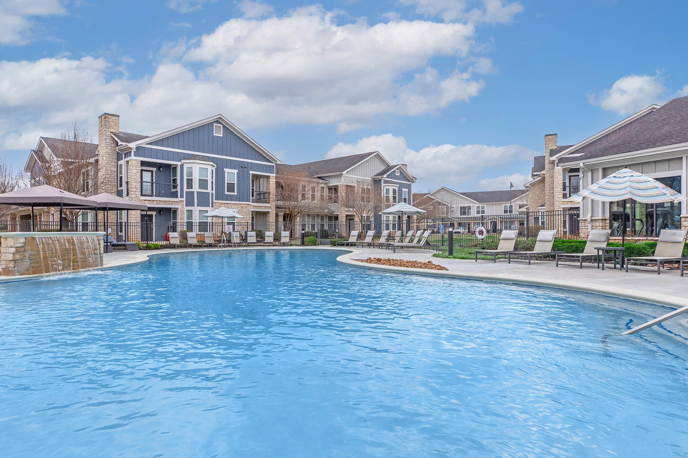 A serene outdoor pool area surrounded by modern apartment buildings. The pool features clear blue water, lounge chairs, and shaded umbrellas. Lush green grass and trees are visible in the background under a partly cloudy sky, creating a relaxing atmosphere for residents and visitors.