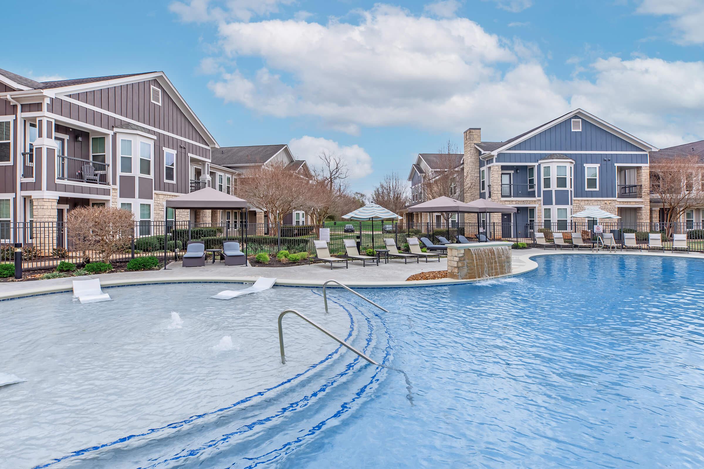 A modern apartment complex pool area featuring a large sparkling blue pool with gentle waves, lounge chairs, and shaded umbrellas. Surrounding greenery and landscaped areas add to the inviting atmosphere under a bright blue sky with fluffy clouds.