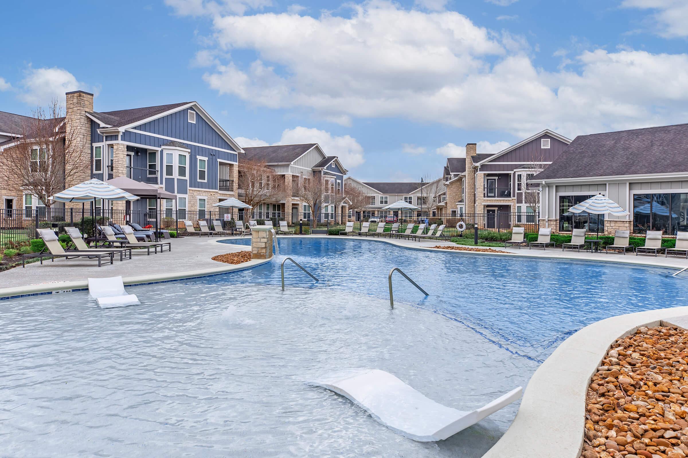 A well-maintained outdoor swimming pool surrounded by lounge chairs and umbrellas, with stylish apartment buildings in the background. The pool features a modern design, including a sloped entry and a decorative stone area. The sky is partly cloudy, enhancing the inviting atmosphere.