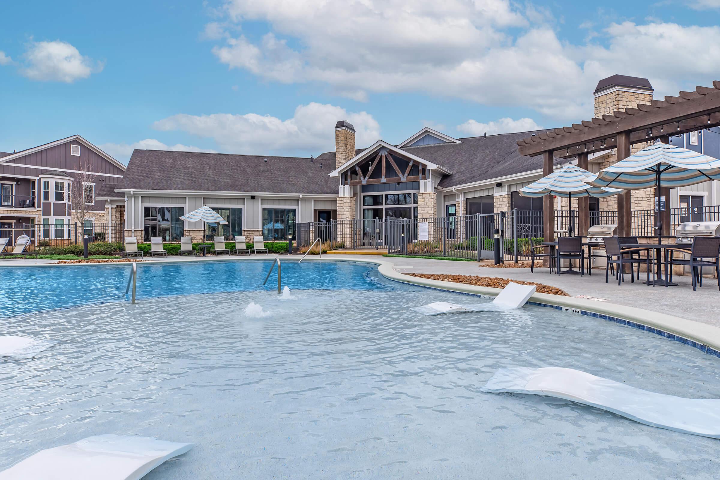 A modern swimming pool with lounge chairs and umbrellas, surrounded by a landscaped area. In the background, there's a stylish clubhouse with large windows and a stone facade. The sky is partly cloudy, creating a relaxing outdoor atmosphere.