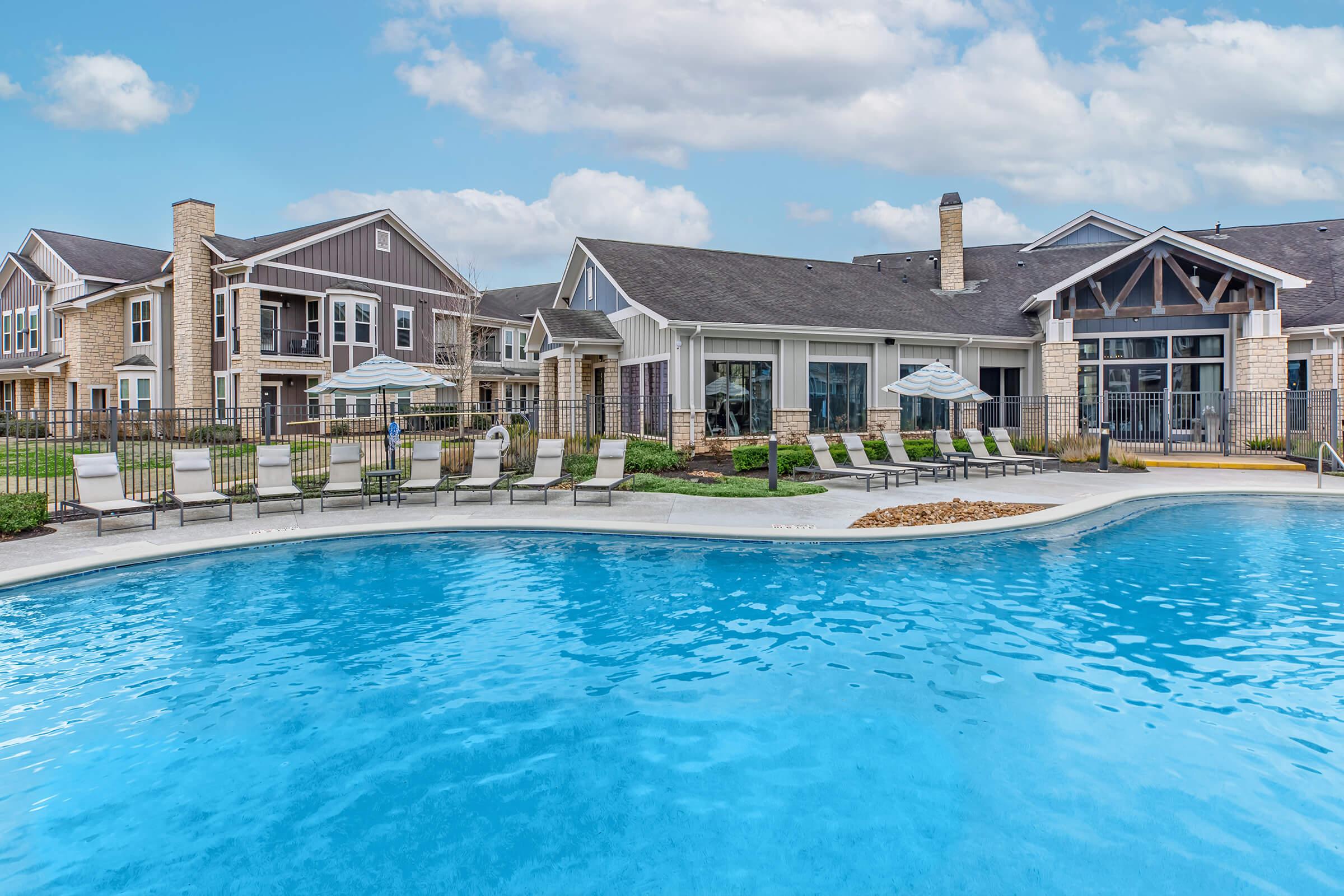 A clear blue swimming pool in an outdoor area surrounded by lounge chairs, with modern residential buildings in the background. The sky is bright with scattered clouds, and there are shaded umbrellas by the poolside, providing a relaxing atmosphere for leisure and enjoyment.