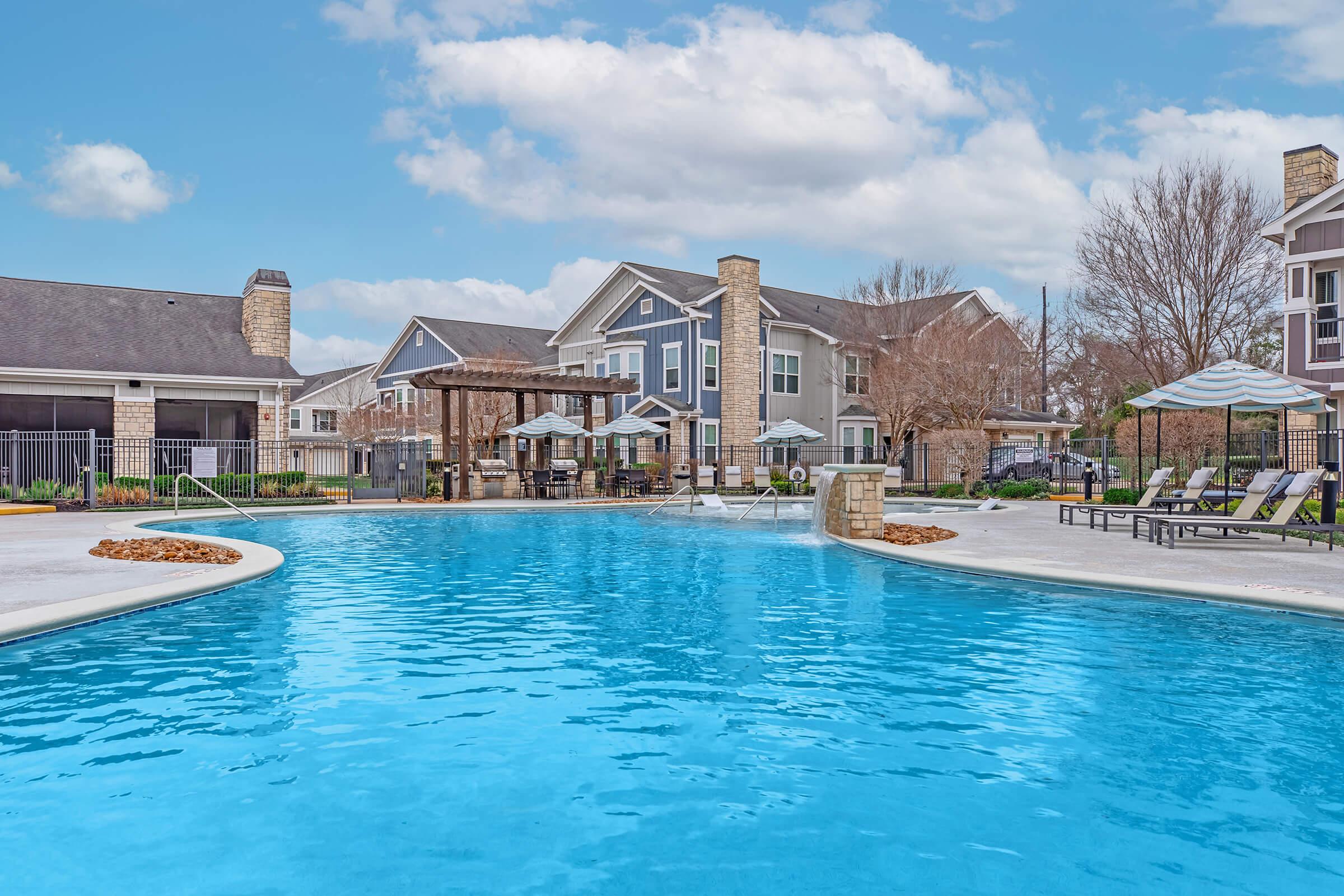 A large swimming pool with clear blue water, surrounded by lounge chairs and shaded seating areas. In the background, there are modern apartments and trees, under a partly cloudy sky. The setting is well-maintained and inviting, ideal for relaxation and leisure activities.