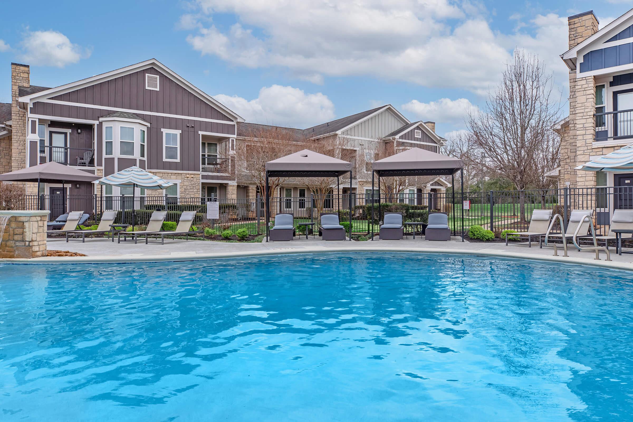 A serene outdoor pool area featuring clear blue water, surrounded by lounge chairs and cabanas. Apartment buildings with a blend of stone and siding are in the background, under a partly cloudy sky, creating a relaxing and inviting atmosphere.