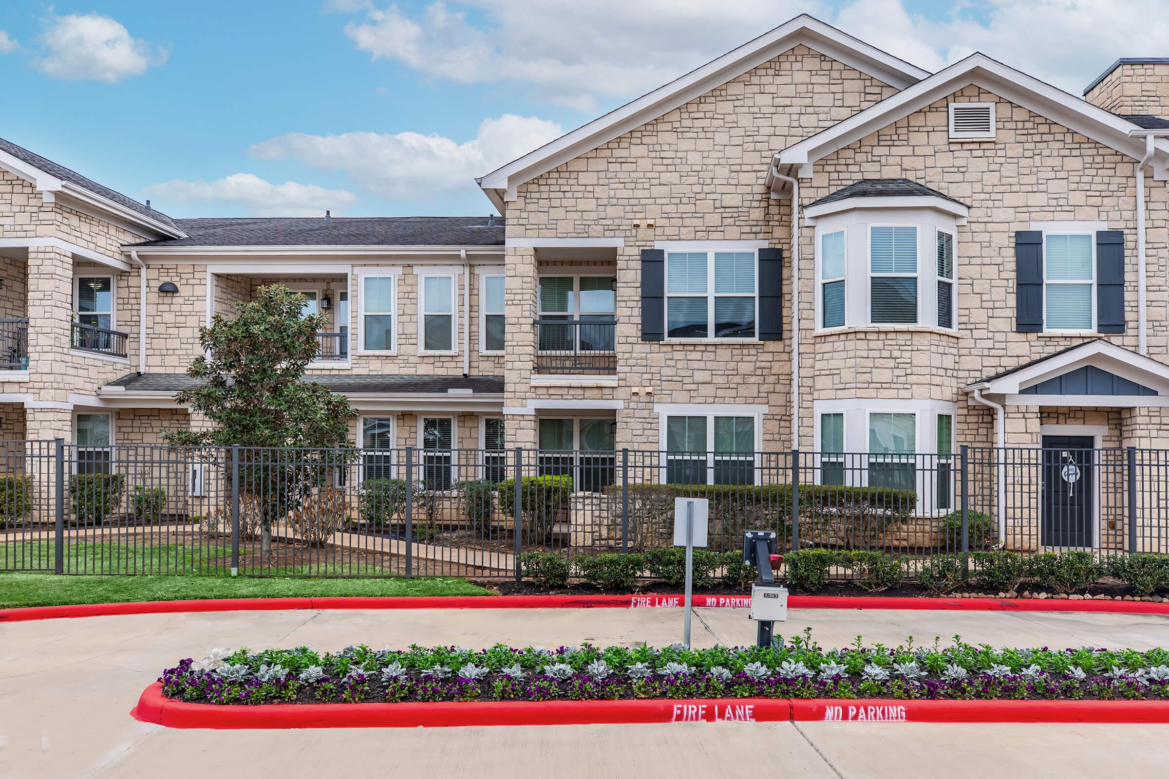 A modern apartment complex with stone exterior and multiple windows. It features a well-maintained landscape with colorful flower beds along a paved area. A fence surrounds the property, and a sign indicates no parking in the fire lane. The sky is partly cloudy.