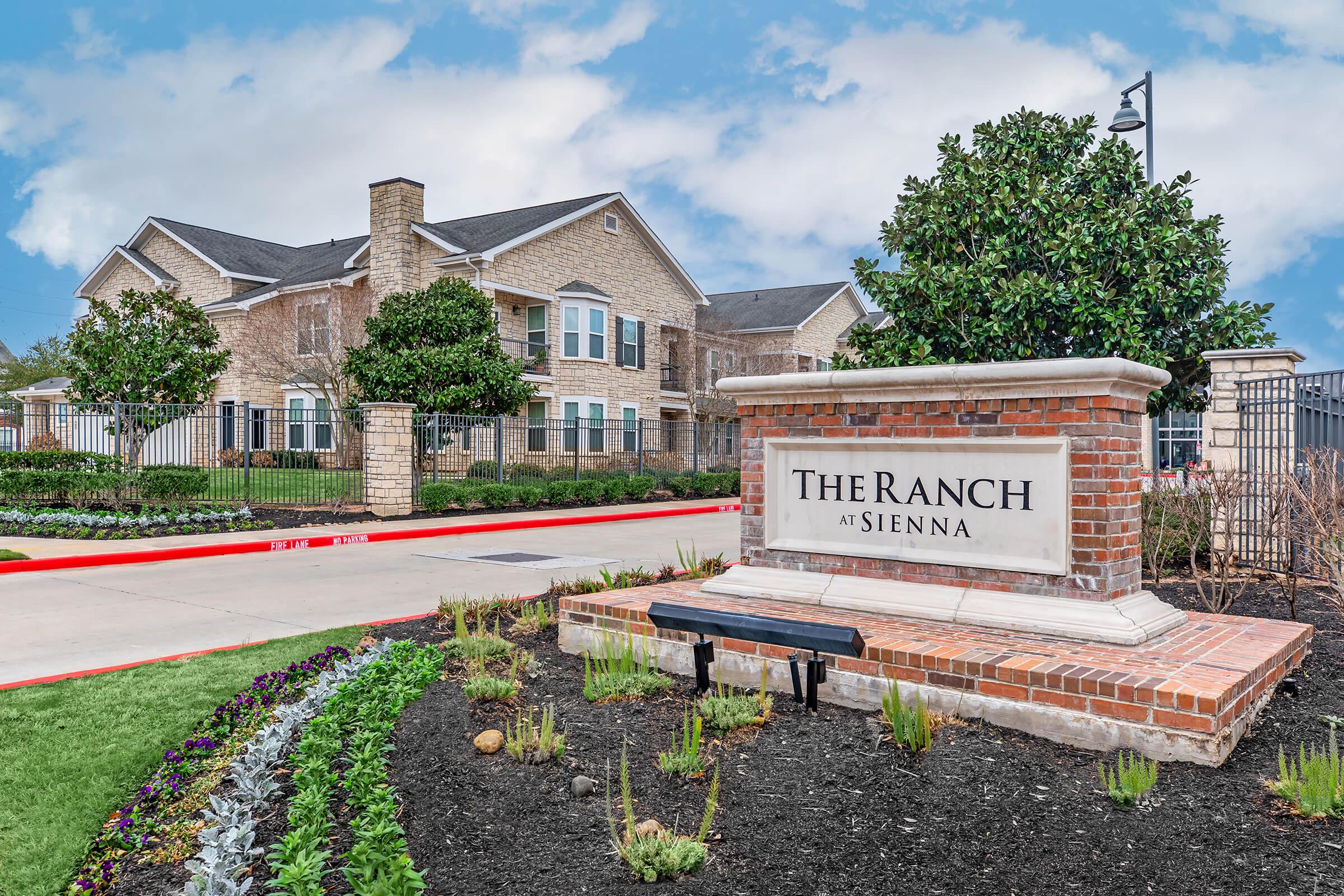 A brick sign reading "The Ranch at Sienna" stands in front of a well-maintained landscaped entrance. The background features a large, modern residential building with multiple windows, surrounded by greenery and flowers, under a partly cloudy sky.