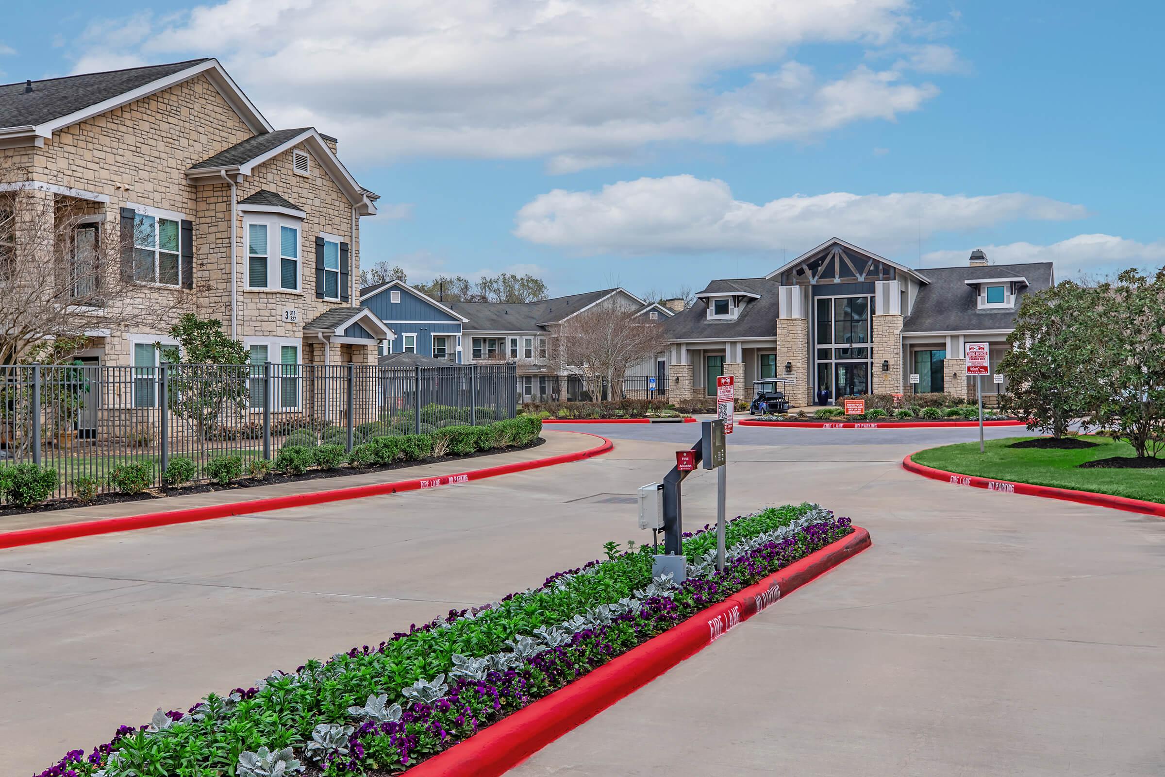 A landscaped entrance to a residential complex featuring two-story brick buildings and a modern main office. A wide driveway with a red curb leads through the property, flanked by vibrant flower beds, under a clear blue sky with a few clouds.