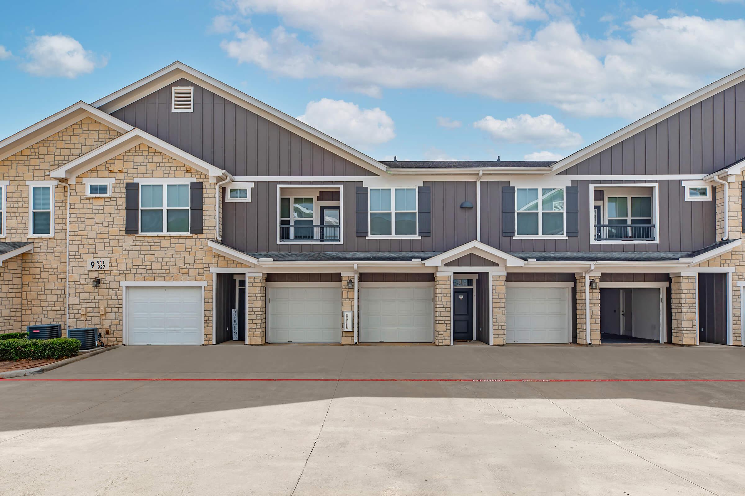 A modern two-story apartment building featuring stone and siding exteriors, with multiple garage doors and balconies. The building is set against a bright blue sky with a few clouds, and a clean concrete driveway in the foreground.