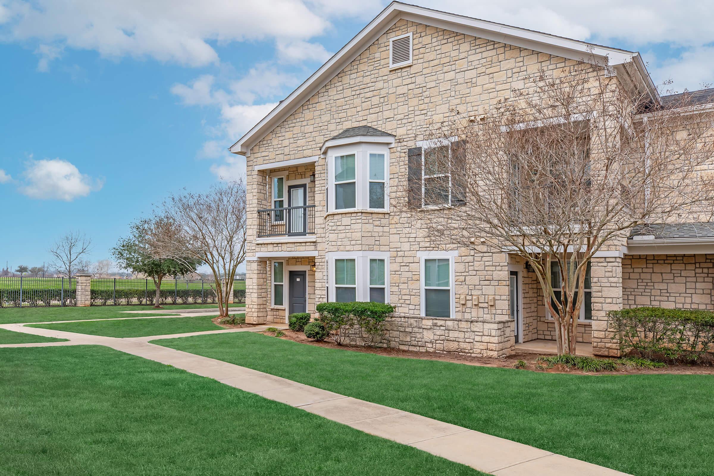 A light-colored stone apartment building with large windows and a well-manicured lawn. There are trees and shrubs lining the pathway leading to the entrance. The sky is partly cloudy, creating a bright and welcoming atmosphere. The building is set against an open field in the background.