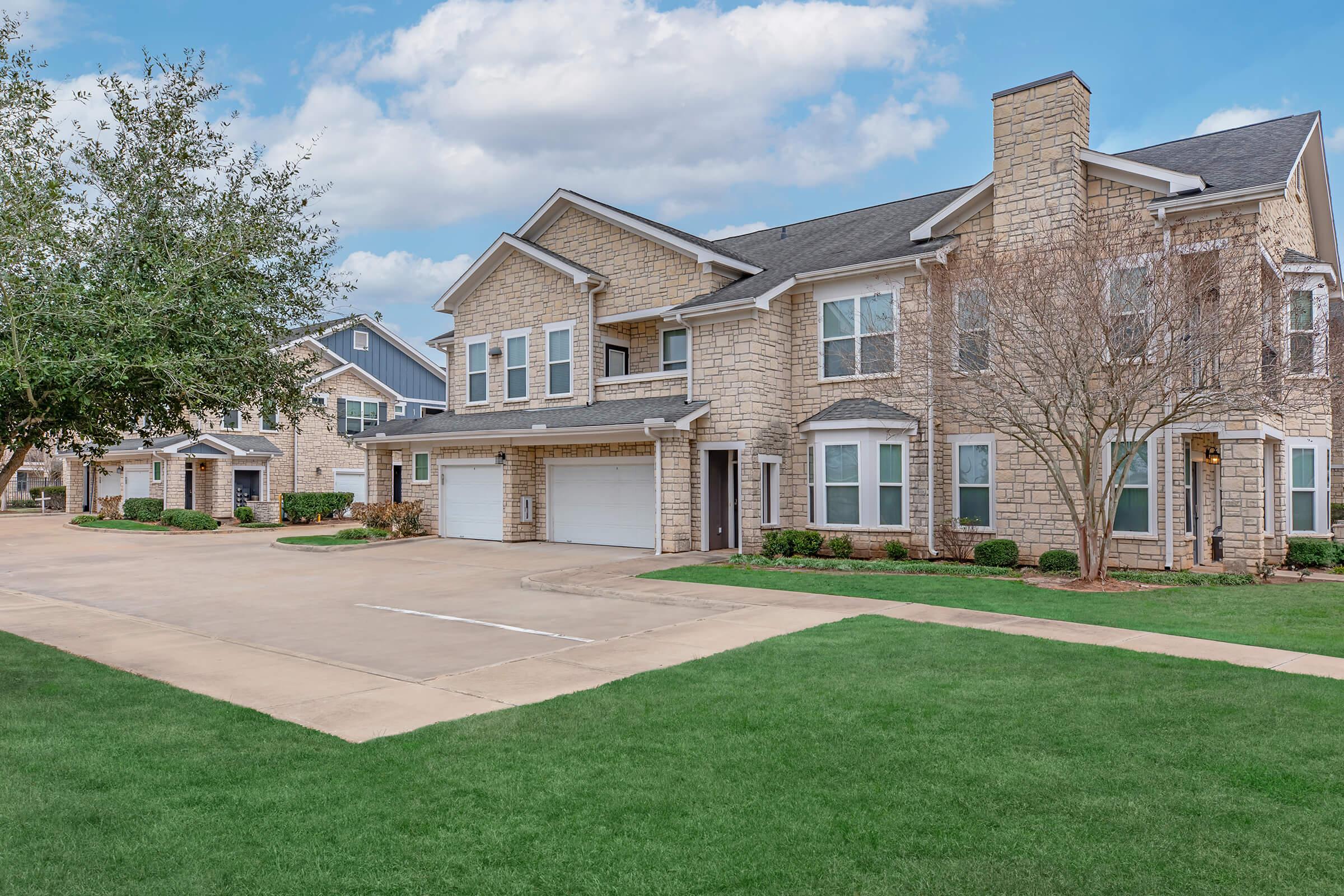 Residential buildings with stone facades and multiple garage doors. The landscaped area features green lawns and trees under a partly cloudy blue sky. The layout shows driveways and well-maintained surroundings, indicating a suburban neighborhood.
