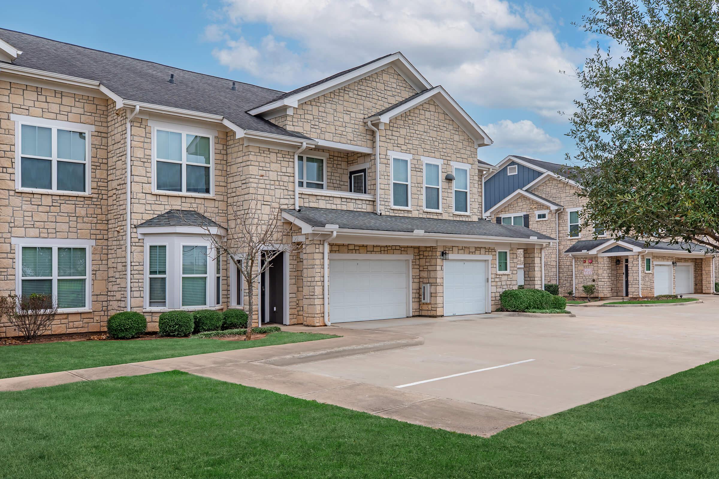Exterior view of a modern two-story apartment building featuring a stone facade, large windows, and a landscaped front yard. Two garage doors are visible, along with a paved driveway and a well-maintained lawn. The sky is partly cloudy, adding to the serene atmosphere of the residential area.