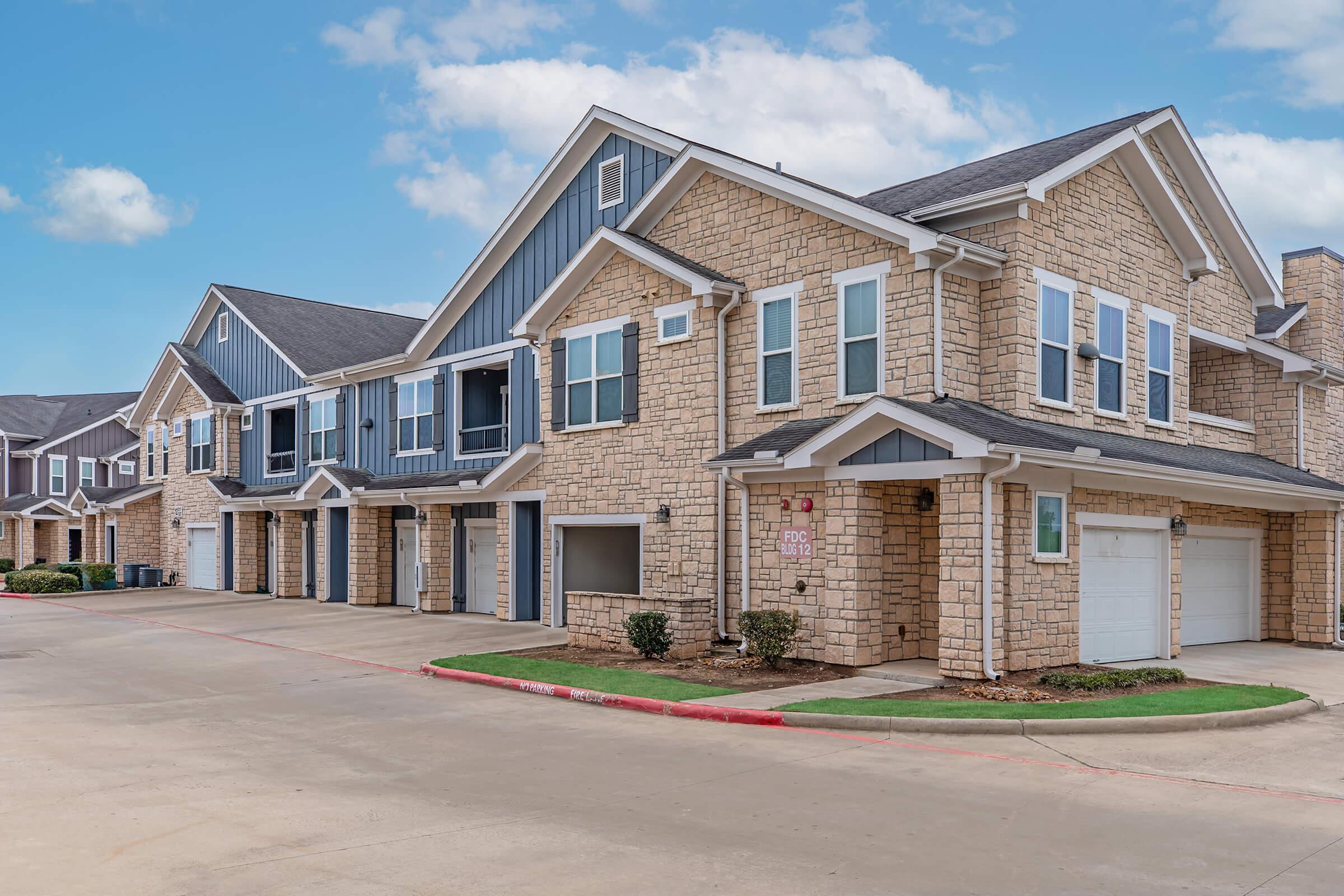 Modern multi-unit residential buildings with stone and siding exteriors, featuring multiple garages and landscaped walkways. The sky is blue with scattered clouds, and the overall setting appears well-maintained and inviting.