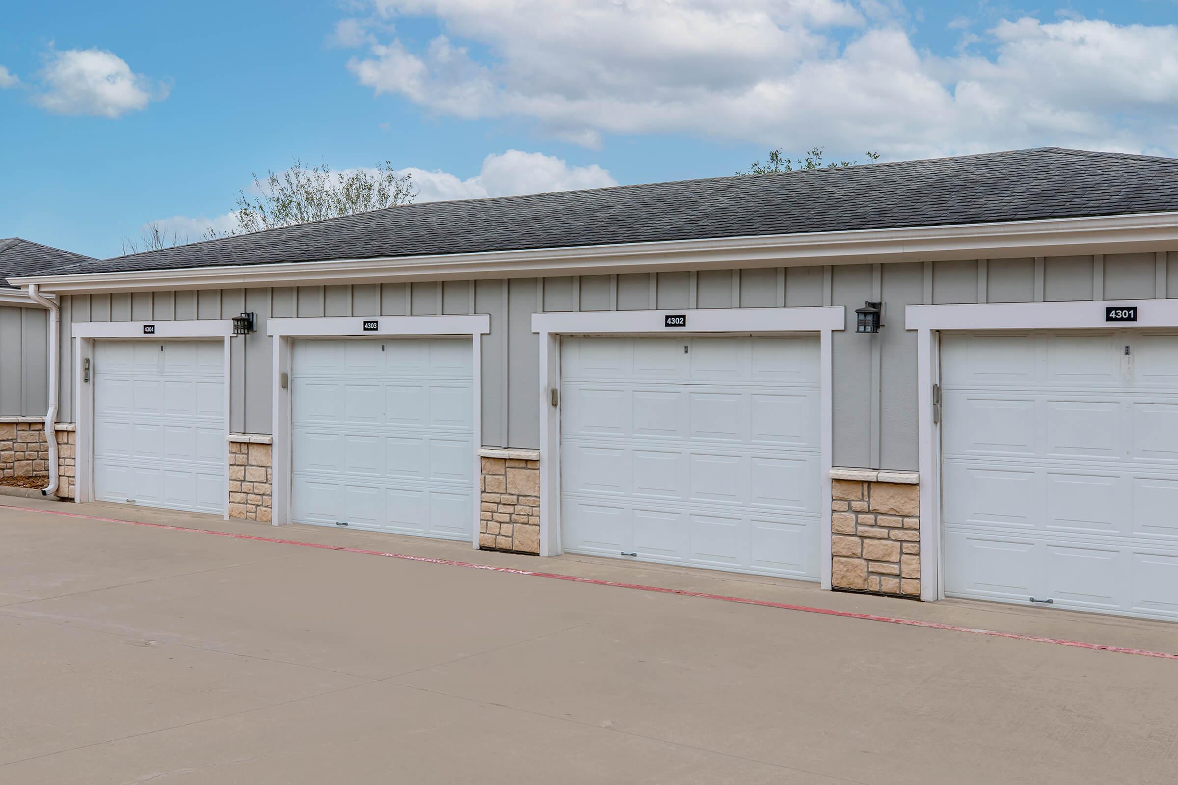 A row of three identical gray garage doors with white paneling and stone accents. Each garage door has a black number above it, and the area is paved with a light-colored concrete surface. The sky is blue with fluffy white clouds in the background.