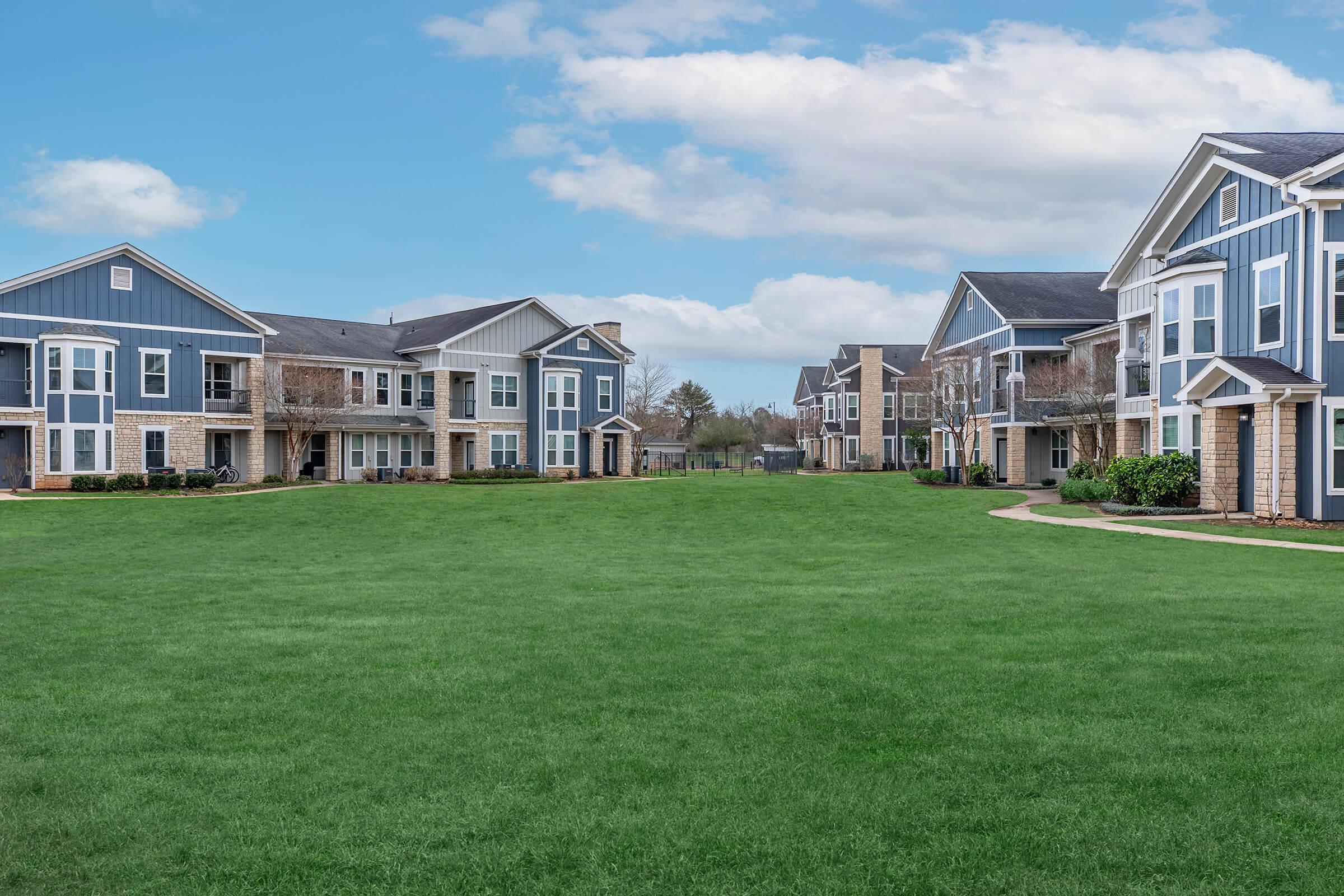 A view of a residential area featuring two-story apartment buildings on either side of a green lawn. The sky is partly cloudy, adding a bright atmosphere to the scene. The buildings have a modern design with blue and beige exteriors. A paved walkway runs through the grassy area, providing access to the apartments.