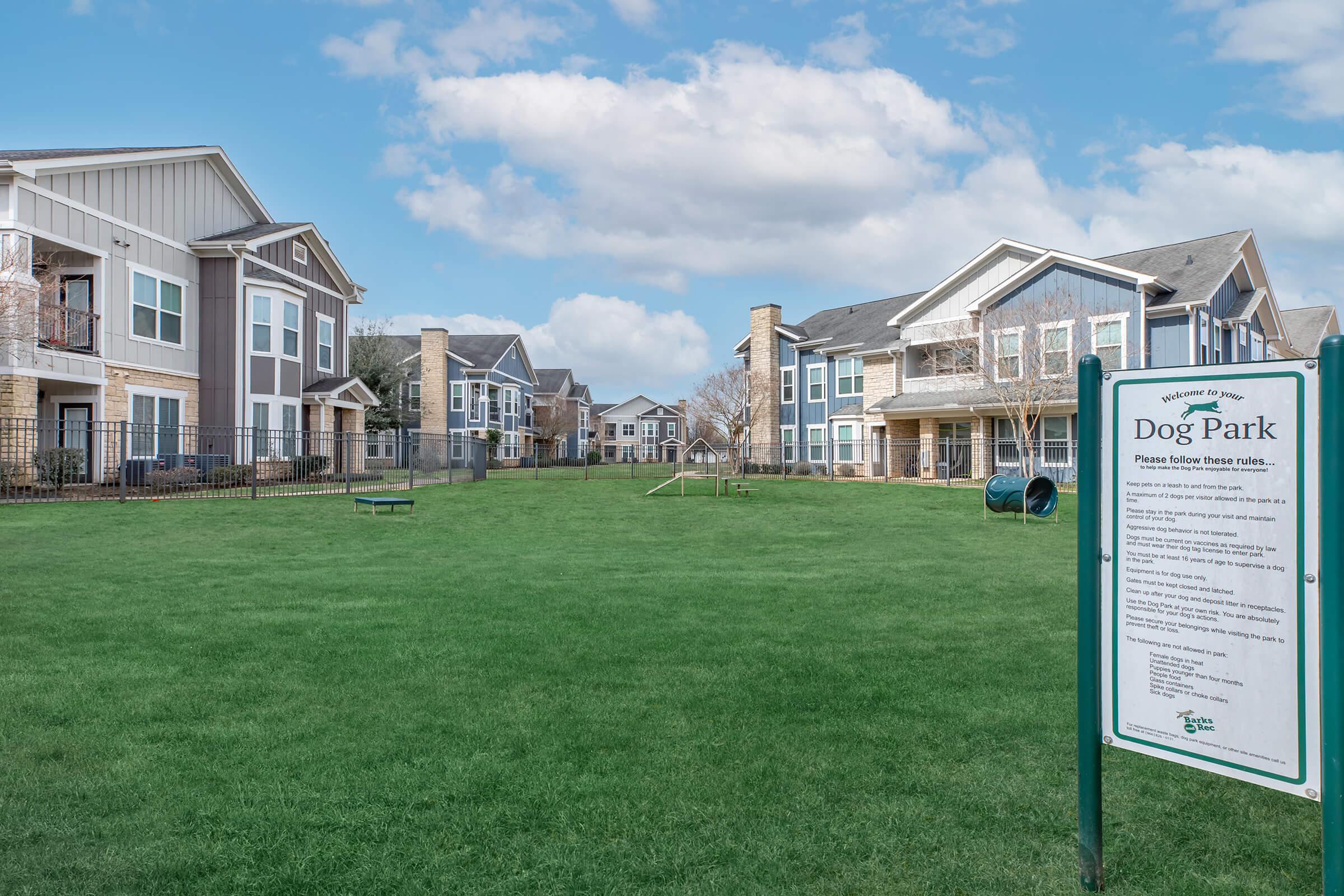 A spacious dog park with green grass, surrounded by residential buildings. A sign detailing park rules is visible in the foreground. The scene features a clear blue sky with a few clouds, providing a pleasant outdoor environment for pets and their owners.