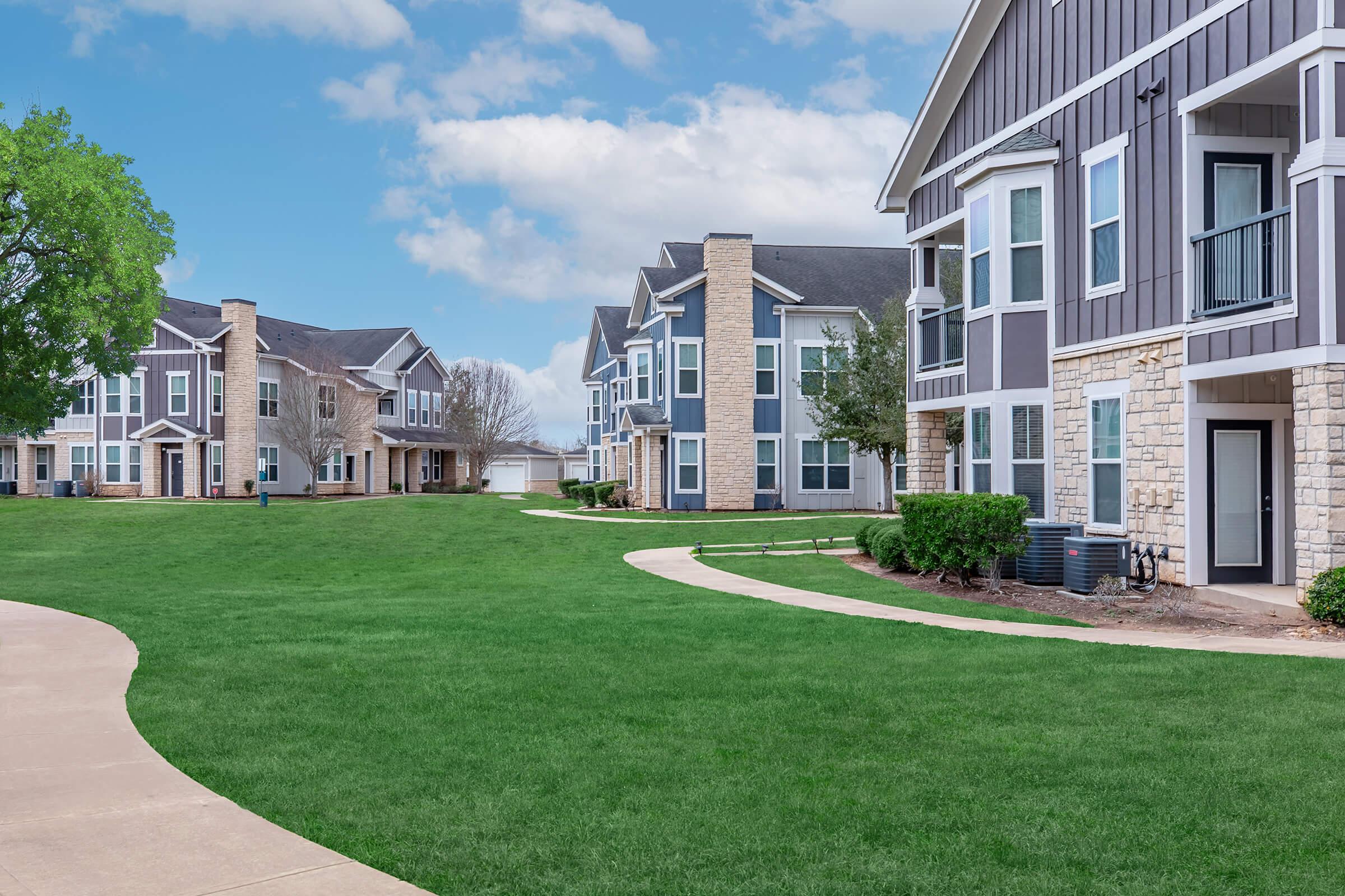 A well-maintained residential area featuring two sets of modern apartment buildings, with manicured green lawns and curved walking paths. The sky is partly cloudy, creating a pleasant atmosphere in the neighborhood.