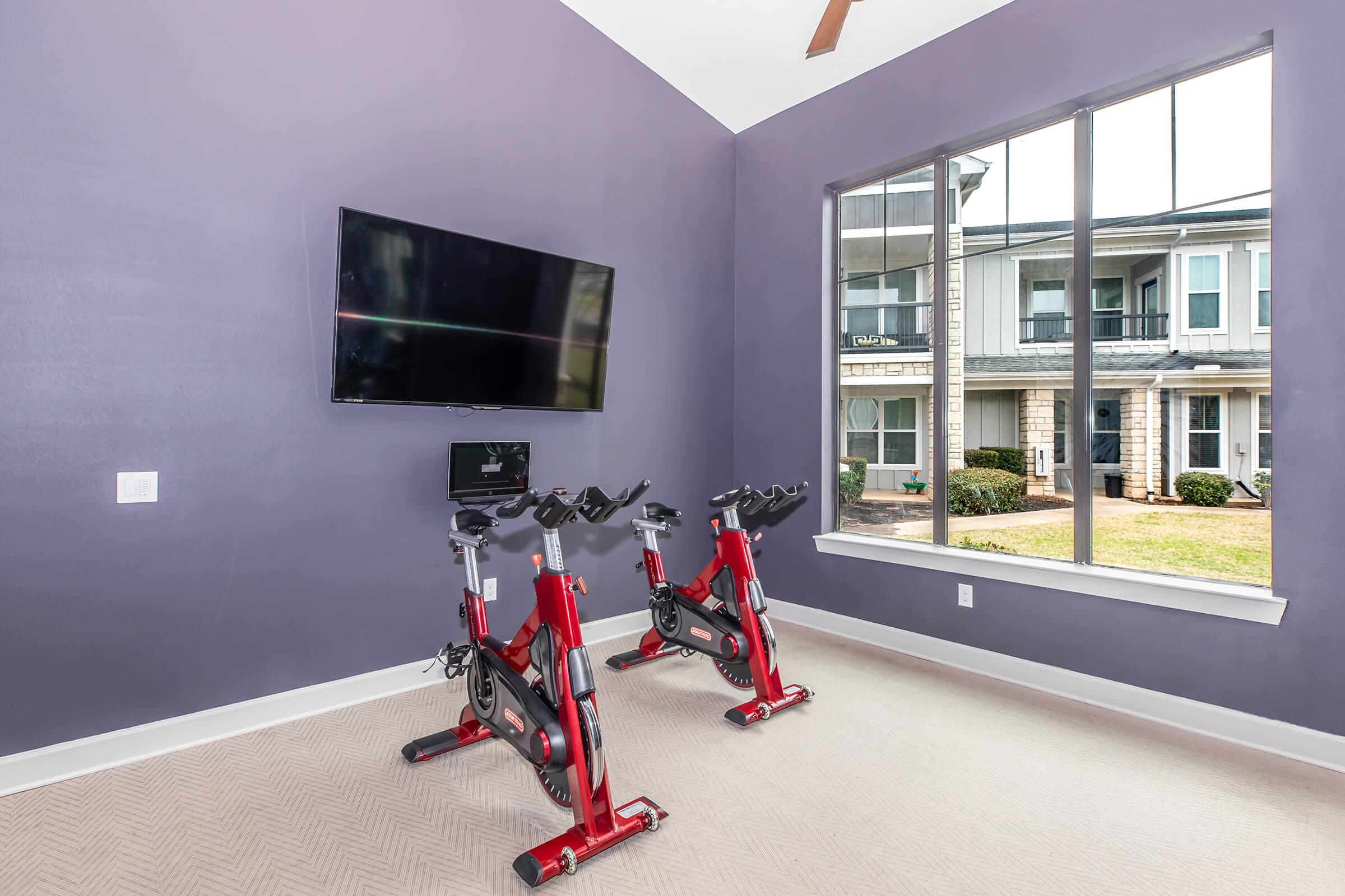 A modern indoor cycling studio featuring two stationary bikes in front of a large window. The walls are painted a rich purple, and there is a wall-mounted flat-screen TV. Natural light streams in from outside, highlighting the comfortable and inviting atmosphere of the space.