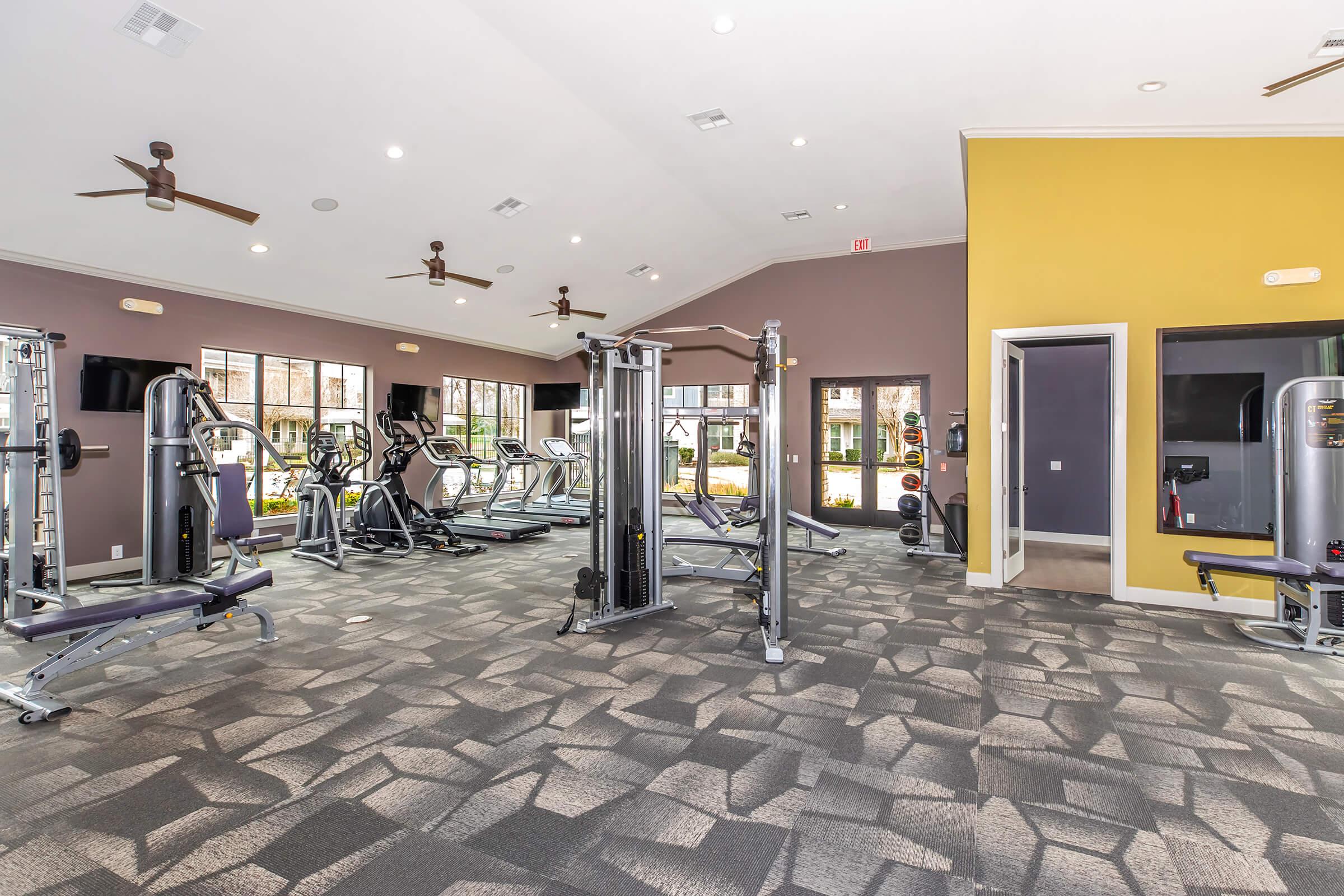 A spacious gym featuring various workout equipment, including cardio machines and weight machines. The room has large windows letting in natural light, decorated with light-colored walls and a contrasting yellow accent wall. Fans are installed on the ceiling for ventilation.