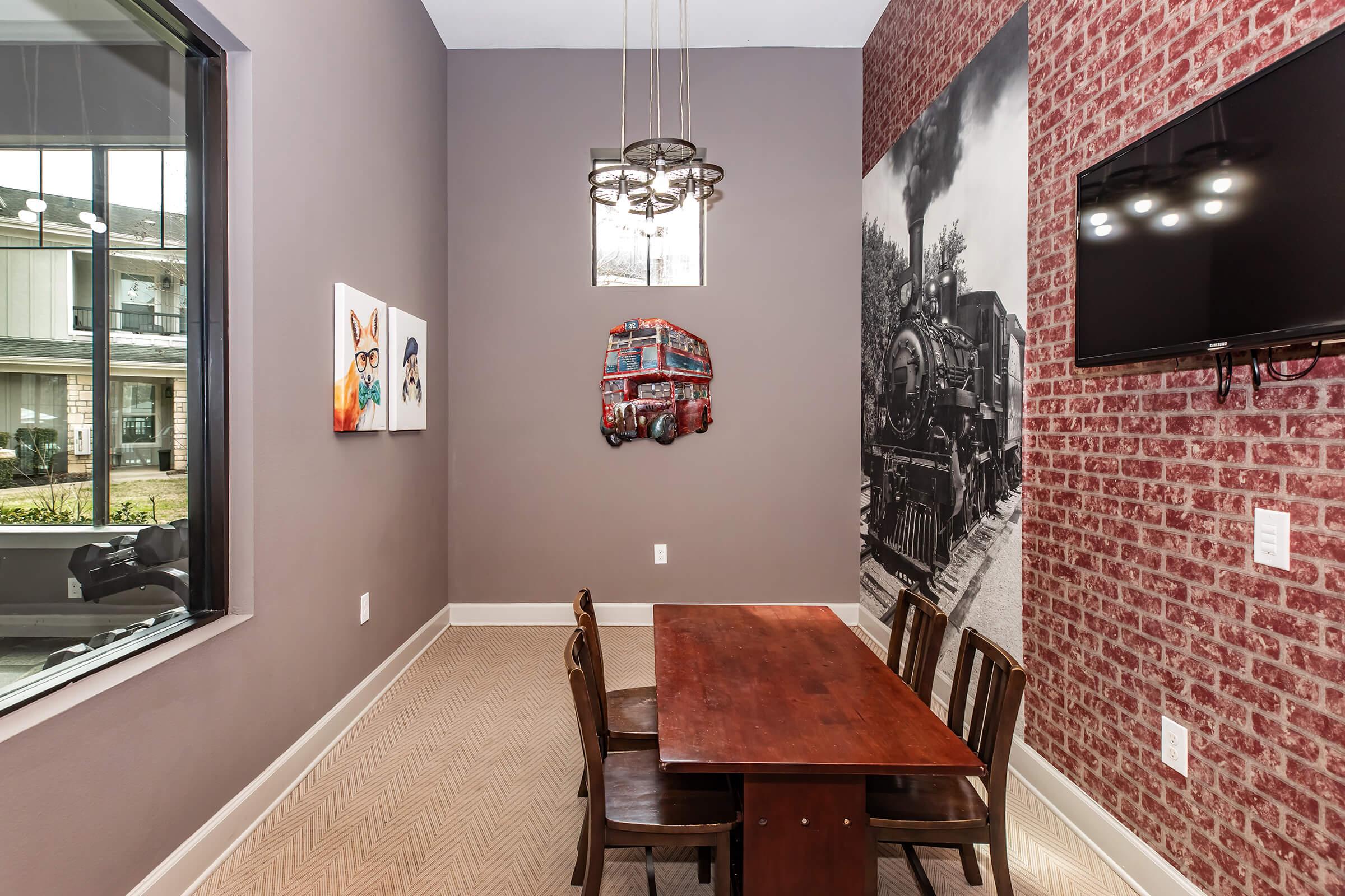 A modern interior space featuring a wooden dining table with four chairs, a large window with natural light, a decorative wall with red brick and an antique train mural, and two colorful art pieces on the adjacent wall. A stylish light fixture hangs from the ceiling above the table.