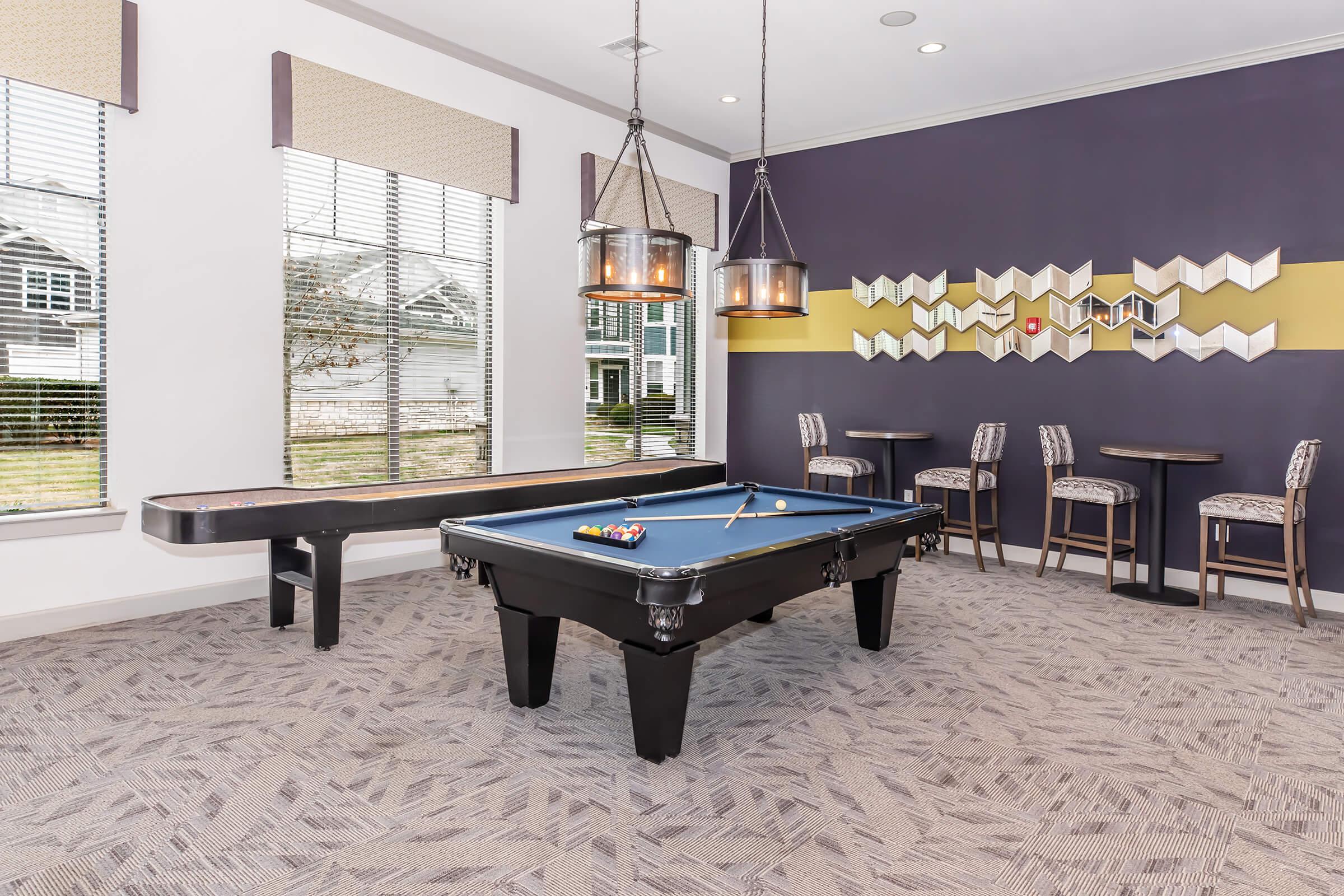 A well-lit game room featuring a blue felt pool table, a wooden shuffleboard table, and stylish high-top chairs. Large windows allow natural light, and modern light fixtures hang from the ceiling, enhancing the inviting atmosphere. Geometric wall decor adds a contemporary touch.
