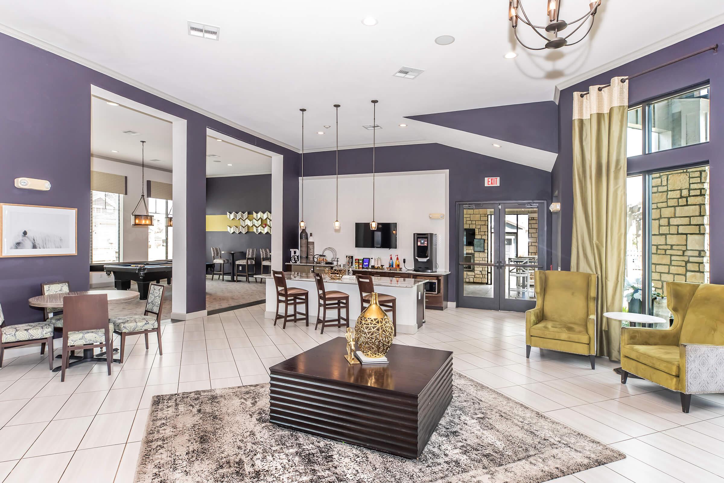 Modern interior of a spacious lounge area featuring a reception desk, seating with green armchairs, and a central coffee table. The walls are painted in purple and beige tones, and there are large windows allowing natural light. Decor includes artwork, a rug, and a billiards table in the background.