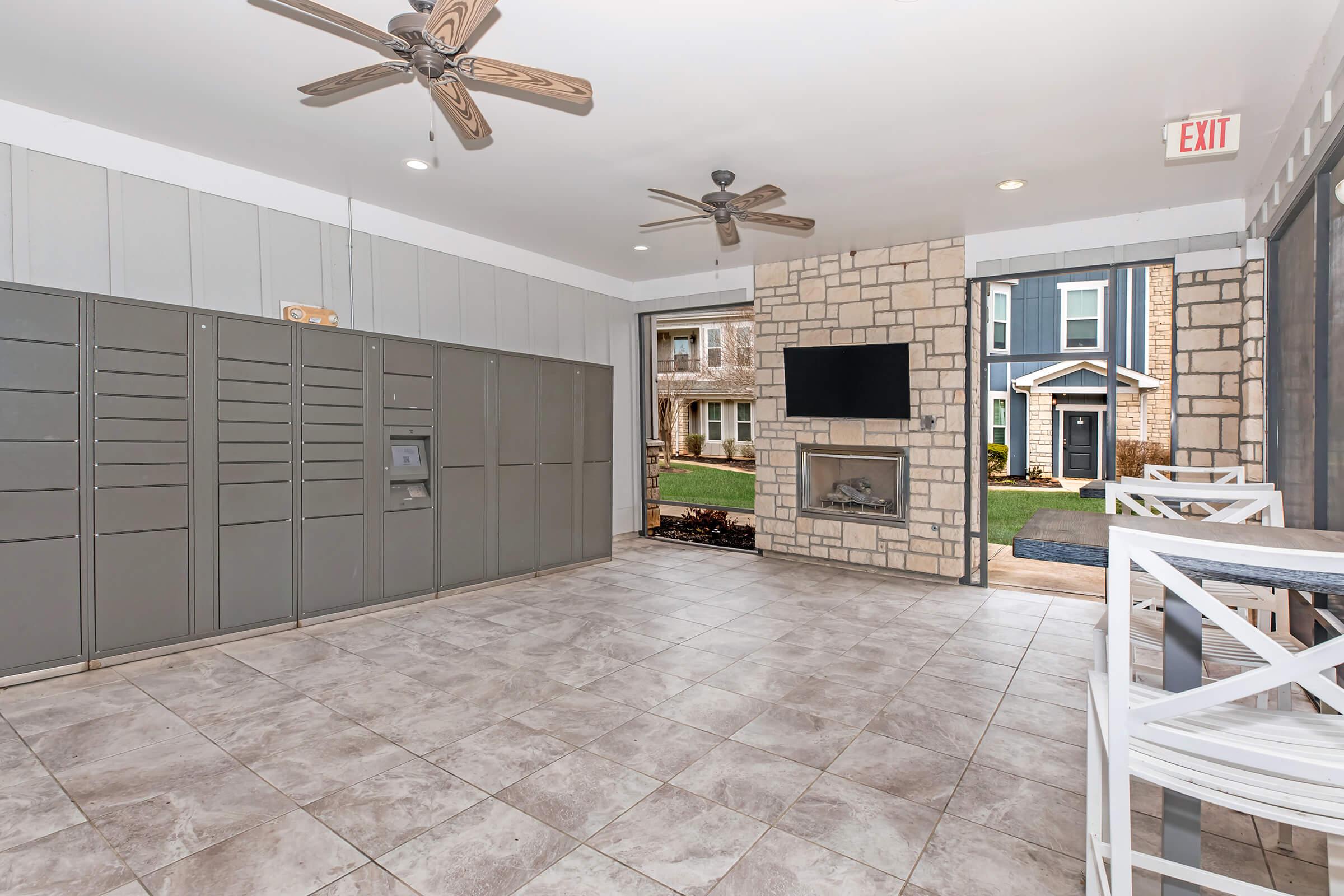Interior of a modern communal space featuring gray mail lockers on one side, a flat-screen TV mounted on a stone wall, and a tile floor. Two ceiling fans are visible. On the far side, there are white outdoor chairs and a view of an outdoor area with greenery and buildings.