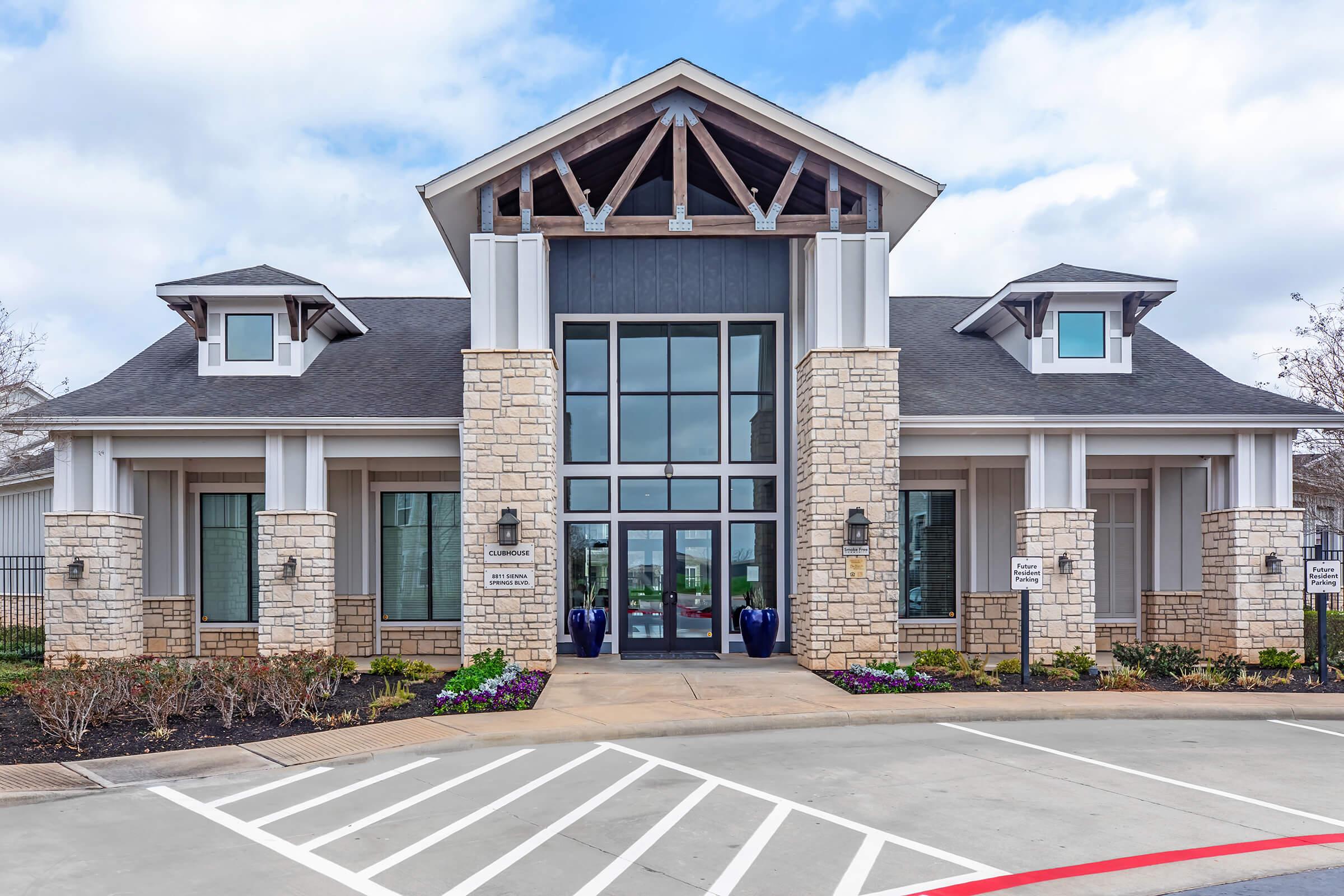 A modern building with a stone and wood facade, featuring large glass doors and multiple large windows. It has a symmetrical design with a peaked roof and decorative elements. The entrance is flanked by blue planters and landscaped areas, set against a cloudy sky. Parking spaces are visible in front.