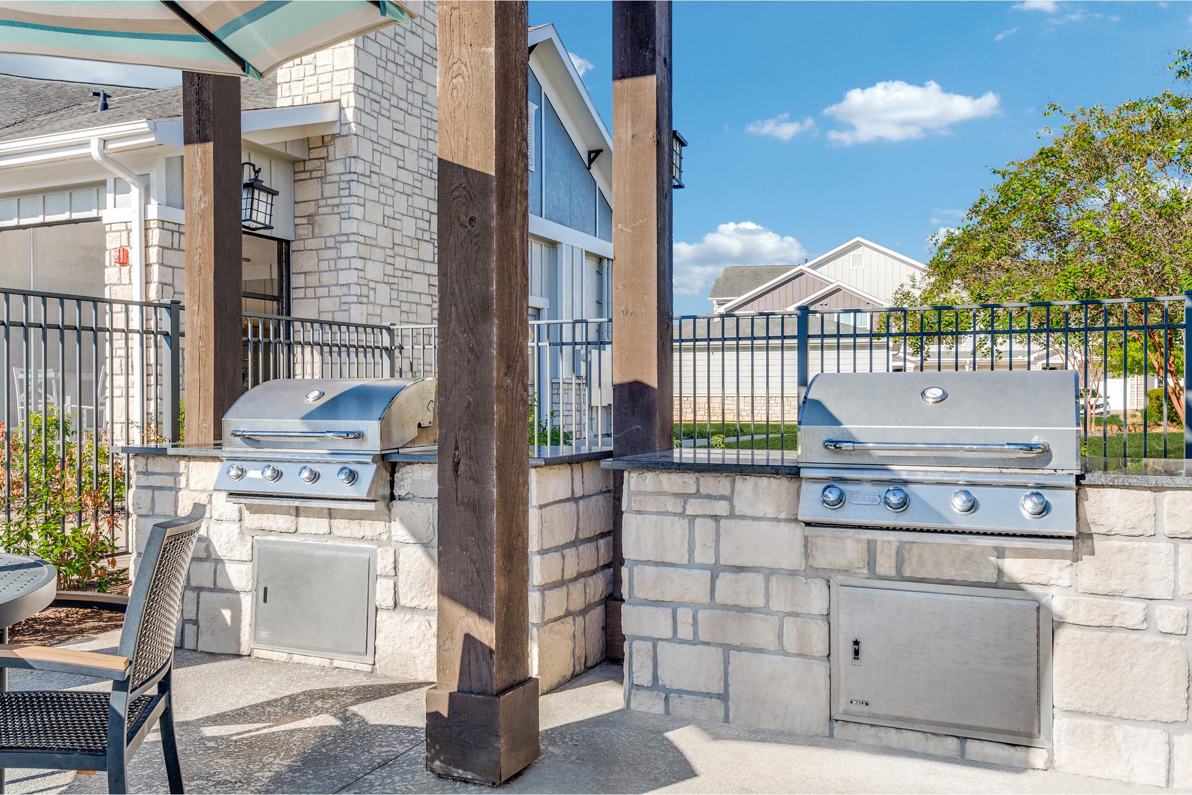 Two stainless steel grills are situated on a stone patio surrounded by a fence. The area features a wooden overhang and is bathed in sunlight, with a blue sky and fluffy white clouds in the background. The grills are flanked by a few chairs at a nearby table, creating an inviting outdoor cooking space.