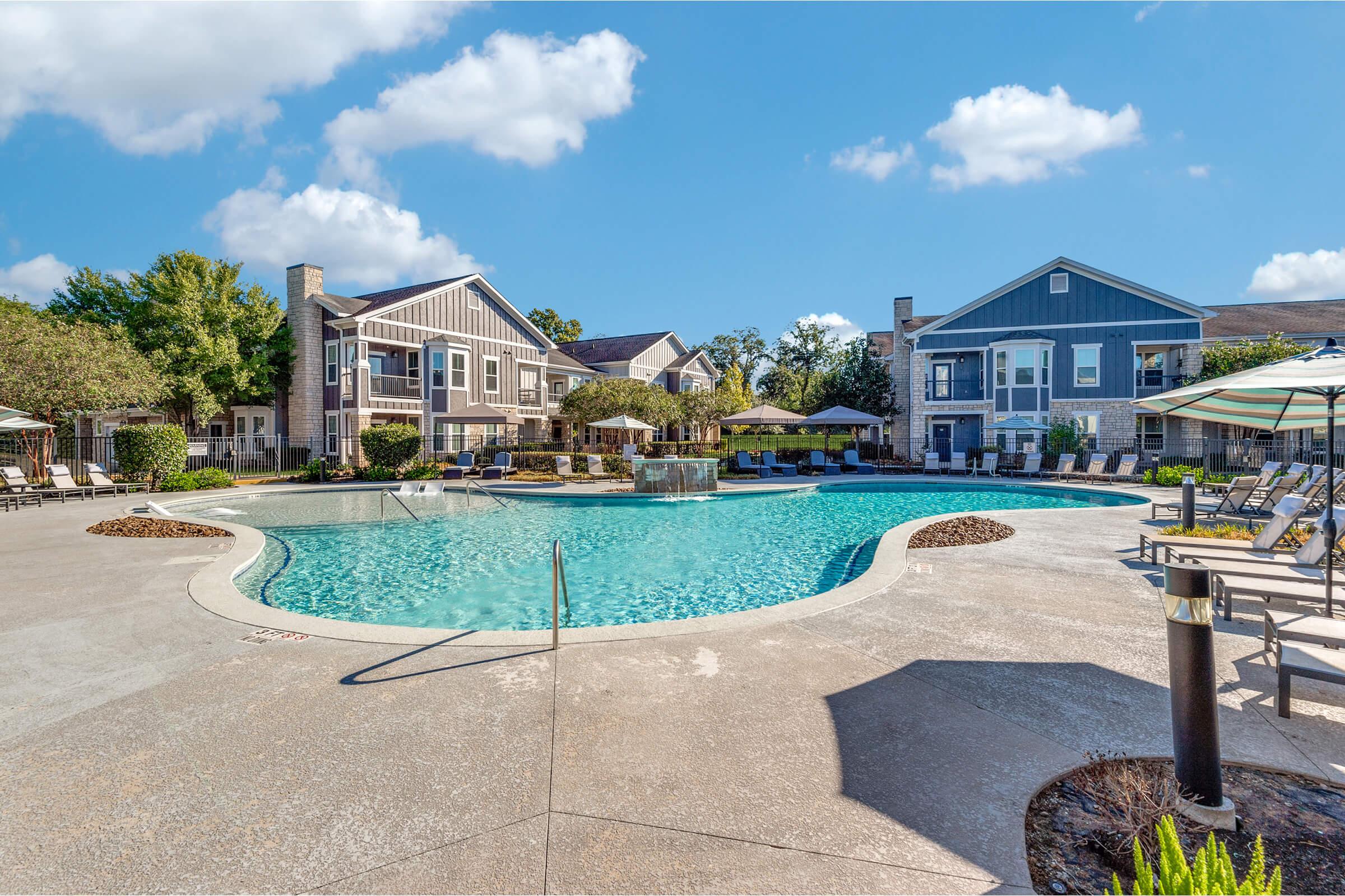 A large, inviting swimming pool surrounded by lounge chairs and umbrellas, with landscaped greenery and modern apartment buildings in the background. The sky is clear with a few clouds, creating a bright, sunny atmosphere. Ideal for relaxation and recreation.