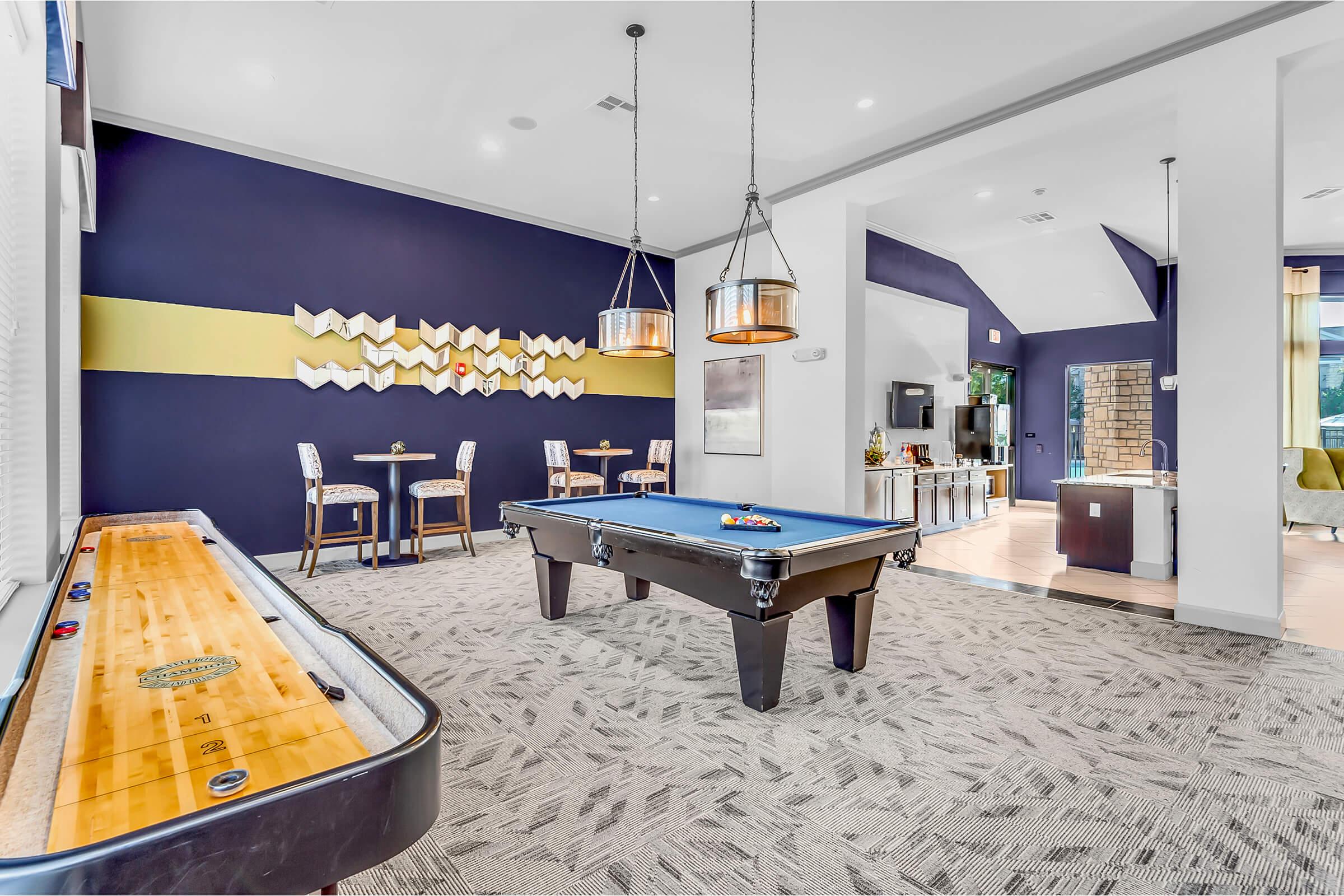 A modern game room featuring a blue billiard table and a shuffleboard table. The walls are painted dark blue with a decorative yellow accent. There are two small tables with chairs in the background, and large windows providing natural light. The flooring is light-colored carpet with a contemporary pattern.