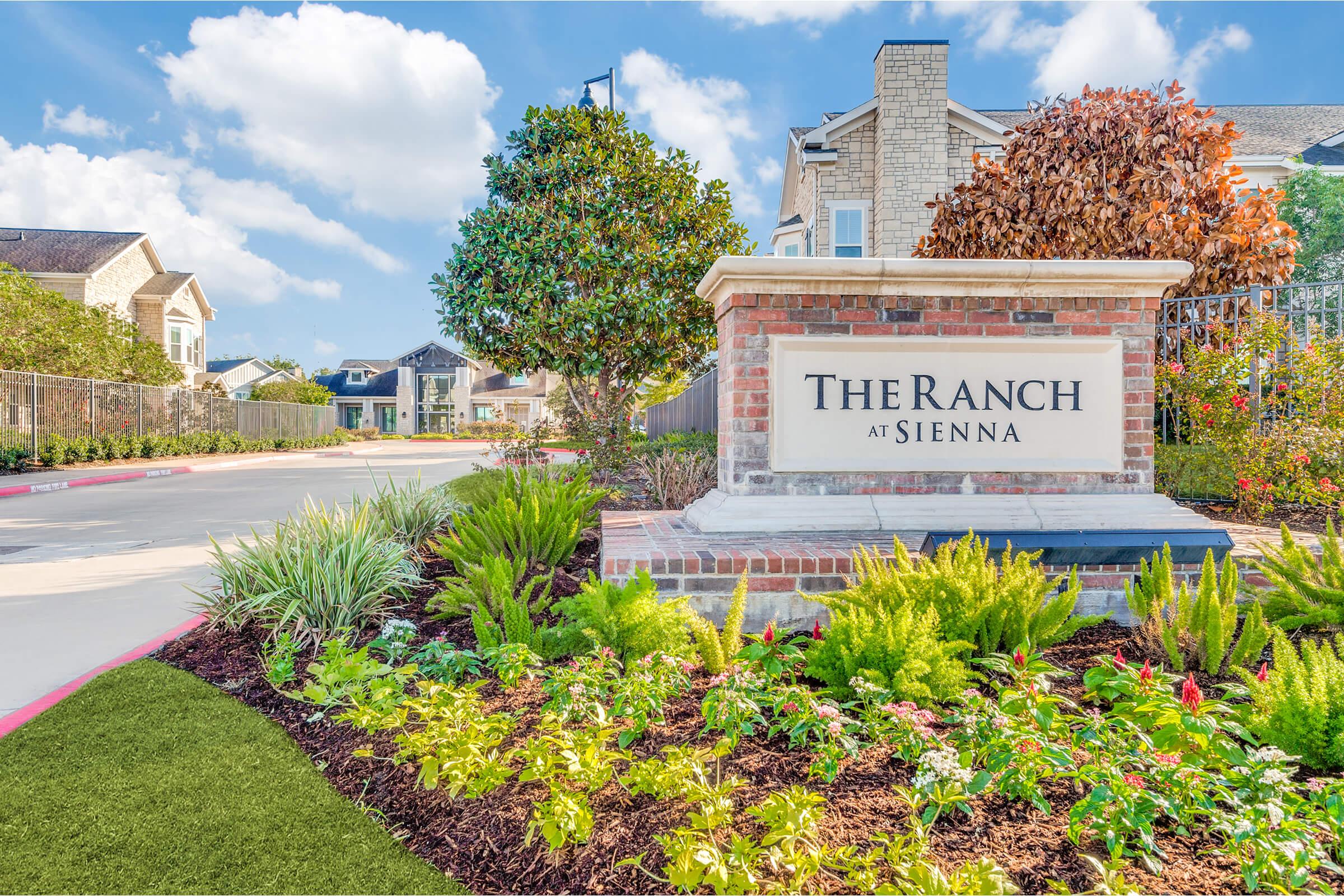 Sign at the entrance of "The Ranch at Sienna," surrounded by well-maintained landscaping, including colorful flowers and shrubs, with a backdrop of residential buildings and a blue sky dotted with clouds. The scene conveys a welcoming atmosphere to the community.