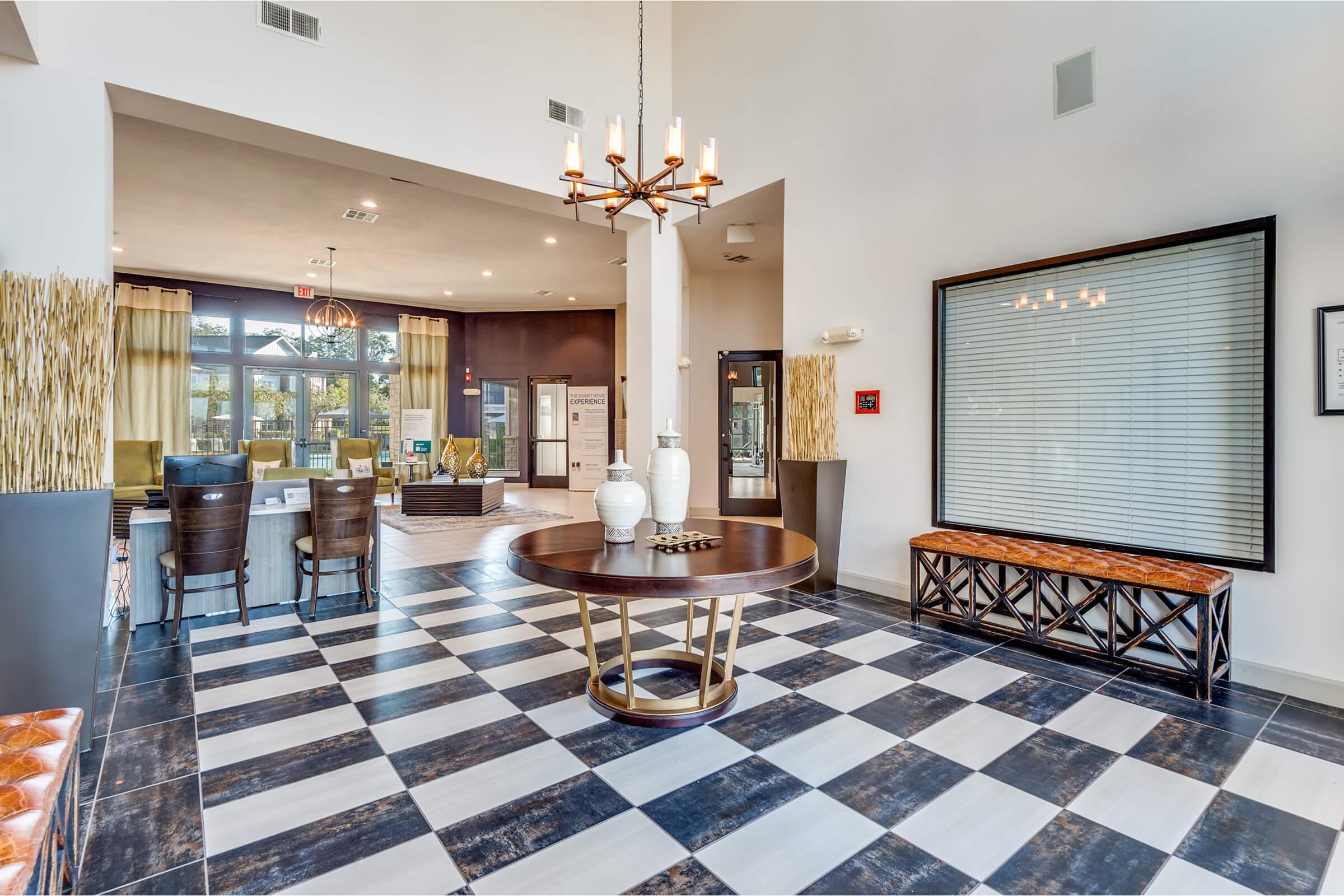 A modern interior space featuring a large round table with decorative vases in the center. The floor has a black and white checkered pattern. In the background, a seating area with green chairs and large windows is visible. A decorative bench and light fixtures add to the contemporary design.