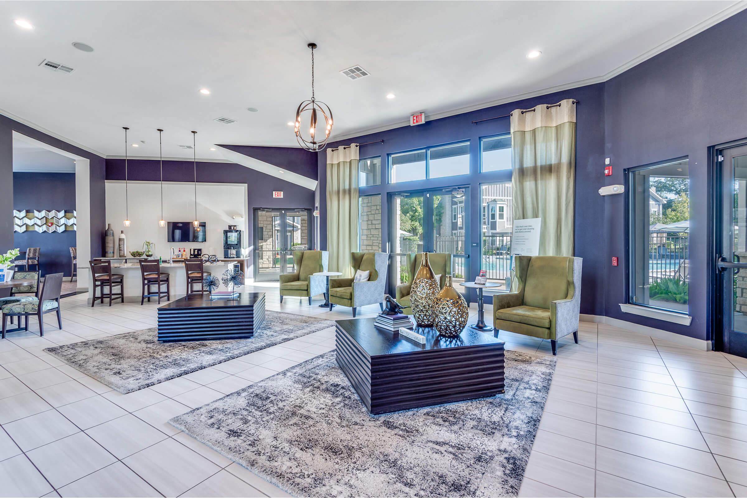 Modern lobby interior featuring stylish seating with green chairs, contemporary tables, and decorative accents. Large windows allow natural light, and a patterned rug adds warmth to the space. The area includes a dining setup and is designed for comfort and elegance.