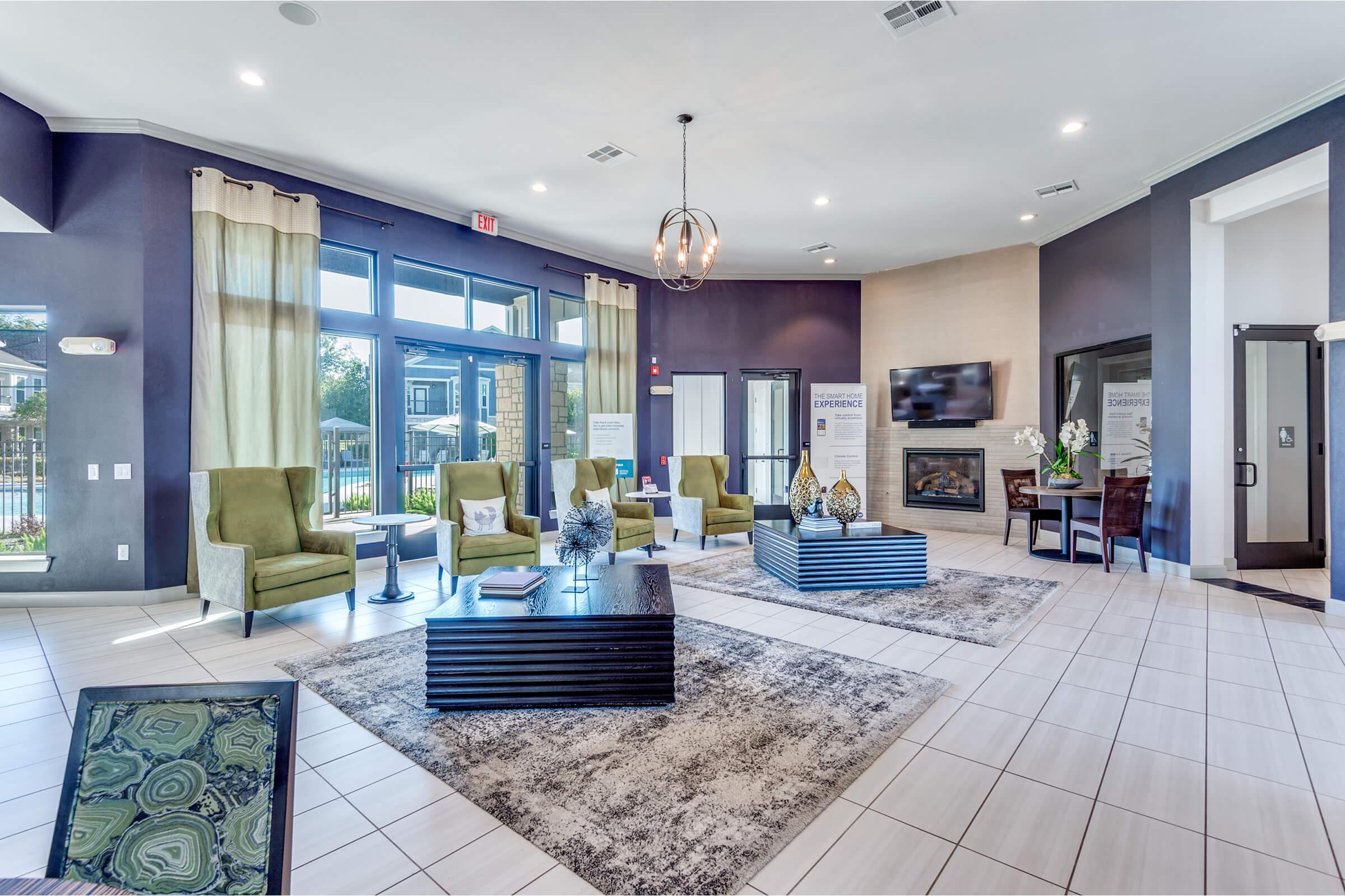A modern lounge area featuring green upholstered chairs, a dark coffee table, and a large area rug. Large windows allow natural light to flood the space, complemented by elegant decor. A flat-screen TV is mounted on the wall, and a fireplace adds warmth to the inviting atmosphere.