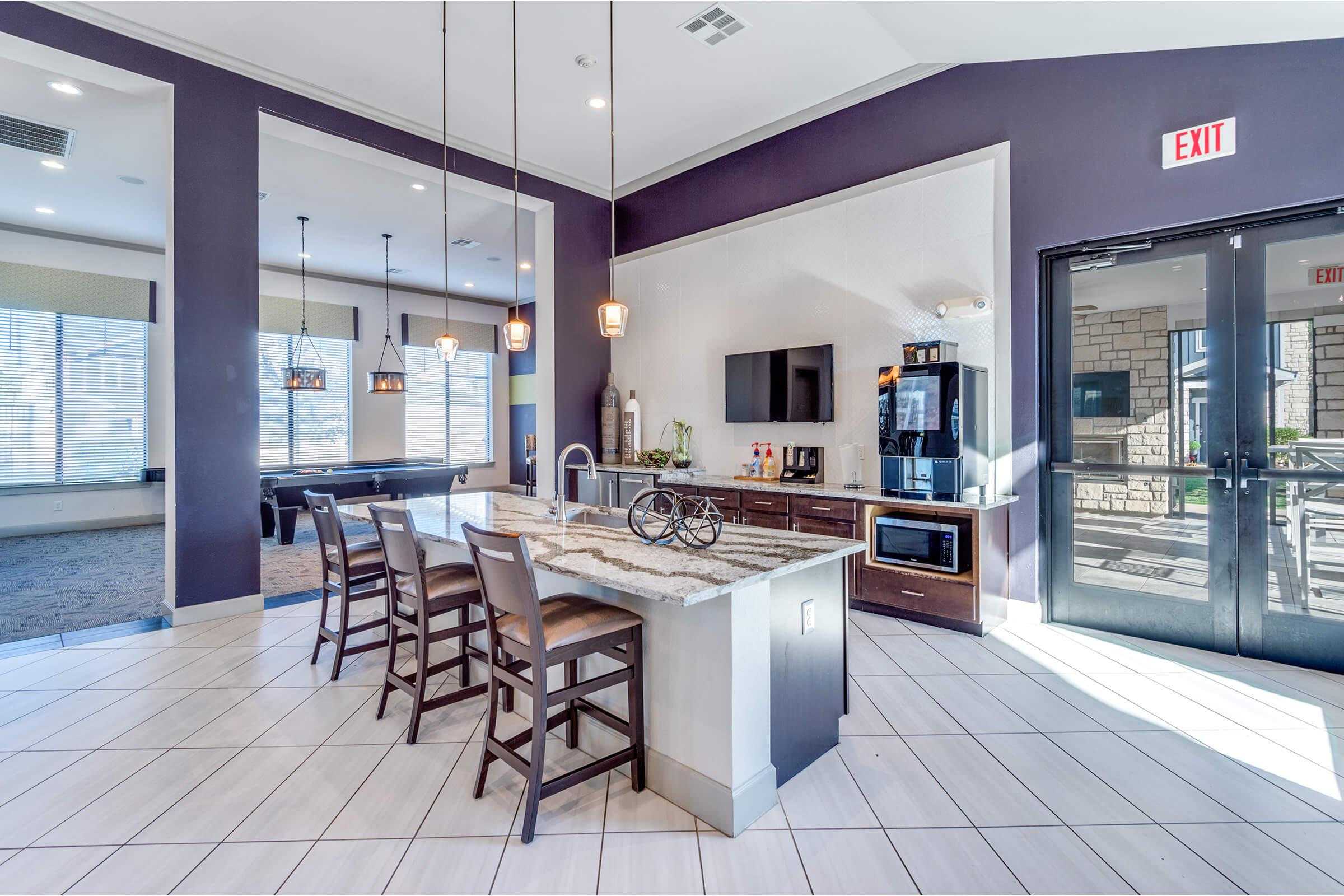 Modern kitchen area featuring a central island with barstools, pendant lighting, and a variety of appliances including a microwave and refrigerator. The space has large windows, light-colored tile flooring, and a door leading outside. There are stylish decorations on the counters and shelves.